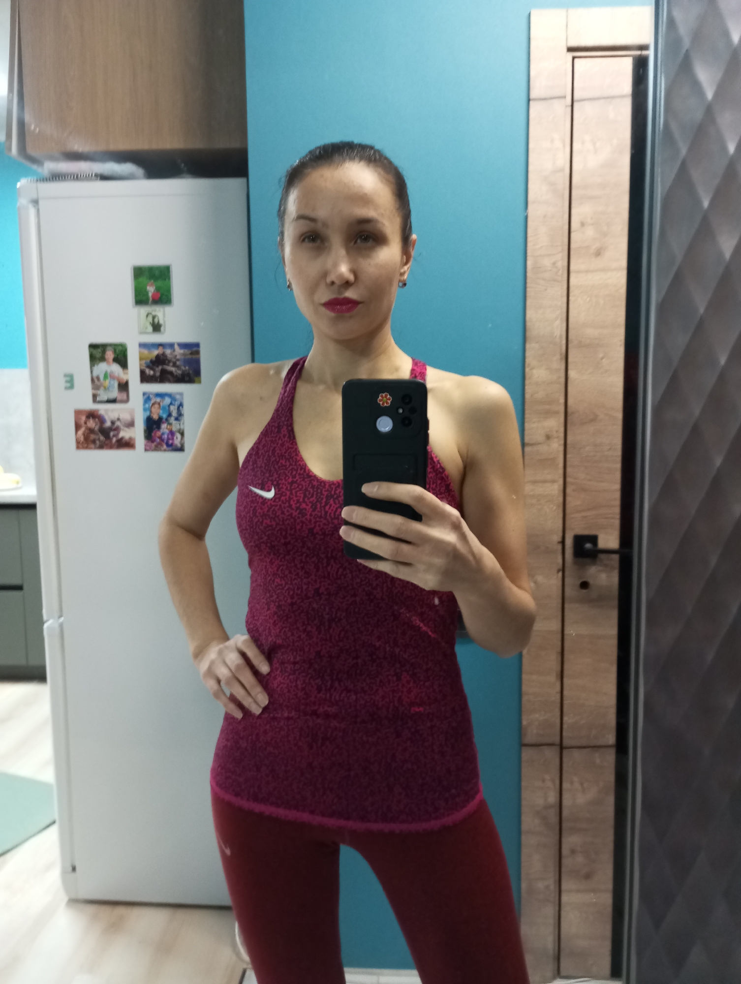 An adult woman is captured in a mirror selfie, holding a black smartphone with her right hand. She is wearing a patterned maroon or deep red Nike tank top with a white Nike logo on the left chest, paired with matching maroon leggings. She stands with her left hand on her hip, looking directly at the camera with a neutral to slightly serious expression.
The setting is an indoor space, likely a kitchen or a room adjacent to one, located in Pervomaysky, Russia. On the left side of the image, a white refrigerator is visible, adorned with several photo magnets depicting individuals and groups of people. Above the refrigerator are light-colored kitchen cabinets. The wall behind the woman and the refrigerator is painted a vibrant blue. To the right, a light brown wooden door frame and a dark, textured door are present. The flooring appears to be light-toned wood. A faint "E" is visible on the side of the refrigerator.