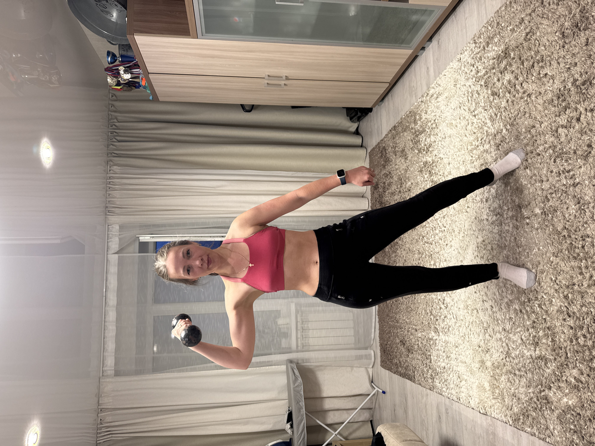 An indoor portrait captures a young woman engaged in an exercise routine within what appears to be a home in Chaikovsky, Russia. The woman, who has light-colored hair pulled back, is positioned slightly to the right, looking directly at the camera with a subtle smile. She is dressed in athletic wear, including a pink sports bra, black leggings, and white socks, and wears a dark-colored smartwatch on her left wrist. In her right hand, she holds a dark dumbbell, indicating she is actively performing a weightlifting or strength training exercise. Her left leg is extended backward, contributing to a dynamic pose.
The background reveals a domestic setting with several details. A large, light-colored, shaggy rug covers a significant portion of the floor, which is otherwise light-toned wood. To the right, a modern wooden cabinet or wardrobe with light-colored and frosted glass doors is visible. Perched on top of this cabinet are several small trophies and medals, suggesting personal accomplishments. Behind the woman, light-colored, pleated curtains obscure a window. In the lower-left corner, a white folding rack or stand is partially seen. The ambient lighting suggests it is evening or night, with artificial light sources creating reflections on the ceiling/wall and the cabinet's glass panels. The scene portrays a dedicated fitness moment in a comfortable home environment. No discernible text is visible.
