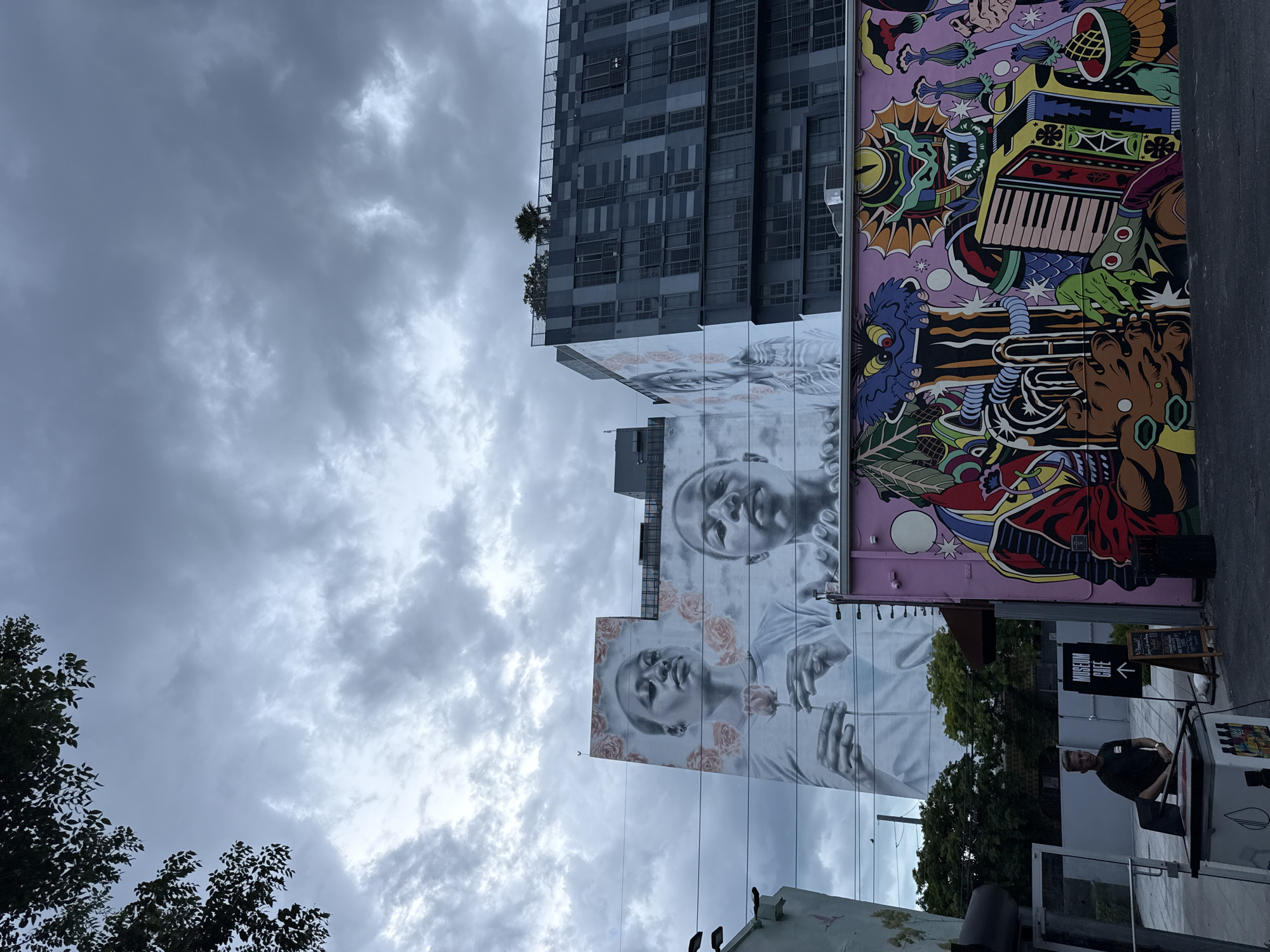The image presents a street scene in Miami, Florida, United States. A tall modern building with a checkerboard facade stands adjacent to a building adorned with vibrant murals. One mural features a large-scale portrait of two figures rendered in grayscale, one above the other, both surrounded by roses. The other mural is a colorful, cartoonish design with musical instruments and abstract elements. Below the murals, there's a "Museum Cafe" sign with an arrow pointing to the left, and a person in a dark shirt stands near a structure that appears to be related to the cafe. The sky above is overcast with a dense cloud cover. Trees with green foliage are visible in the foreground and partially obscure the base of the murals and the cafe area.