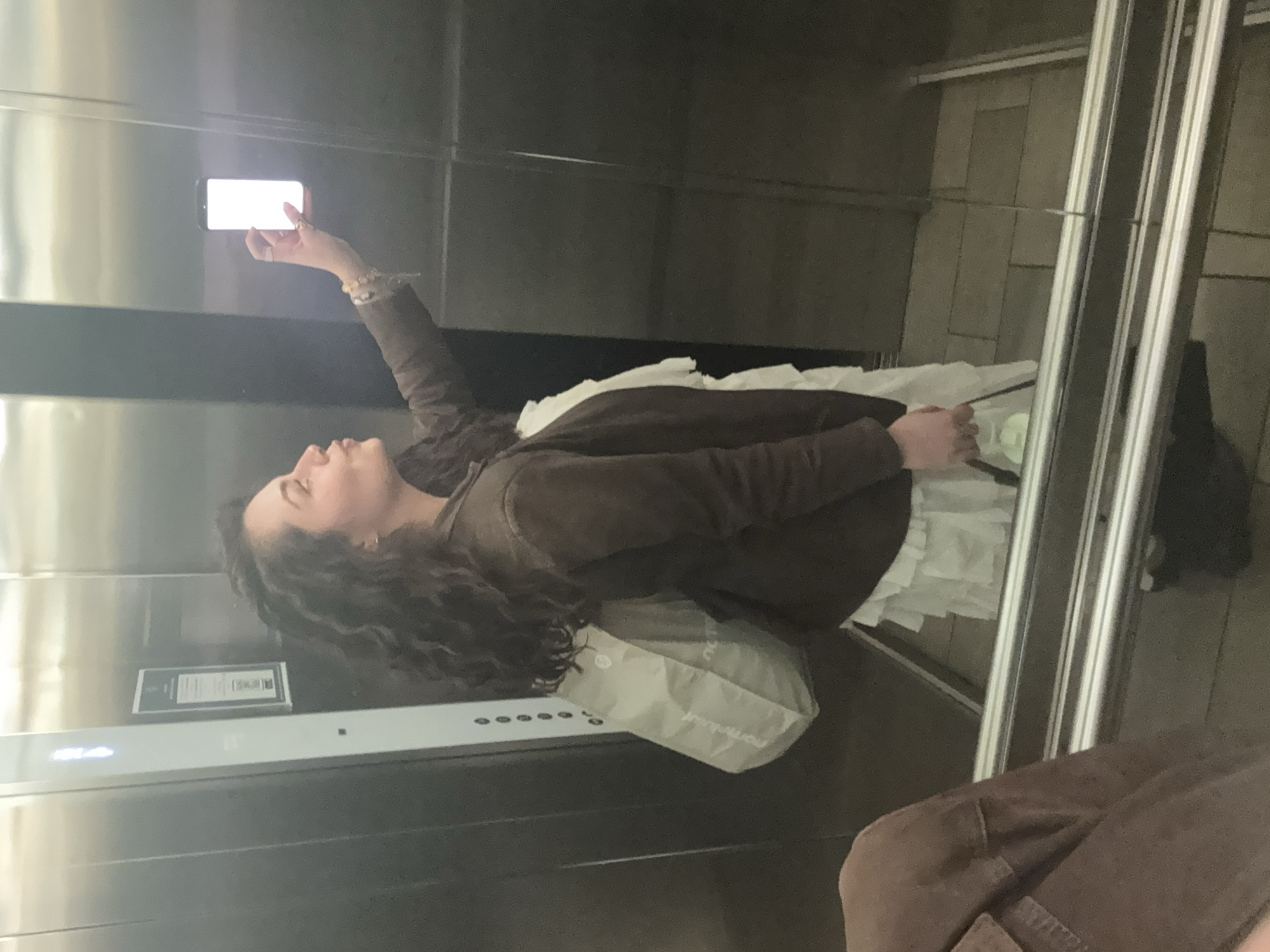 A person with long, dark curly hair is holding a smartphone with a bright screen towards the upper left in an elevator. The person is wearing a brown jacket and is carrying a white bag. A white ruffled garment is also visible in the elevator. The elevator has brushed metal walls and an information panel on the left. There is metal framing to the right of the person, possibly a door frame, with a glimpse of the floor outside the elevator. The location is within the city of Mexico City, Mexico.