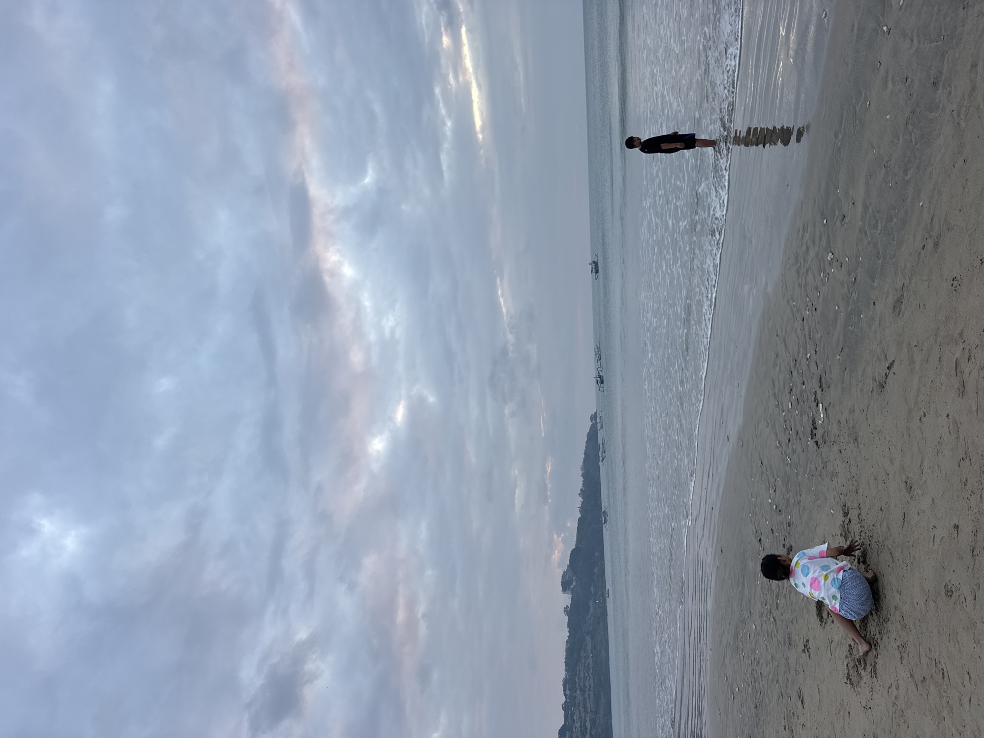 The image depicts a serene beach scene in Kuta Selatan, Indonesia, likely during the late afternoon or early evening, given the soft, diffused light. The sky is overcast with dramatic, textured clouds that hint at a recent or approaching rain shower, yet the overall mood is peaceful.
In the foreground, a young child is crouched on the sandy beach, facing away from the camera. They are wearing a white t-shirt adorned with colorful polka dots and patterned shorts, and appear to be engaged in playing with the sand.
Further back, near the water's edge, a lone figure, possibly a teenager or young adult, stands facing the ocean. They are dressed in dark clothing, including shorts. The calm waves gently lap at the shore, reflecting the muted tones of the sky. In the distance, several outrigger boats are visible, suggesting this is a popular coastal area. The coastline to the left is lined with lush green hills, indicating a tropical setting. The overall impression is one of quiet contemplation and simple enjoyment of the natural environment. There is no visible text in the image.