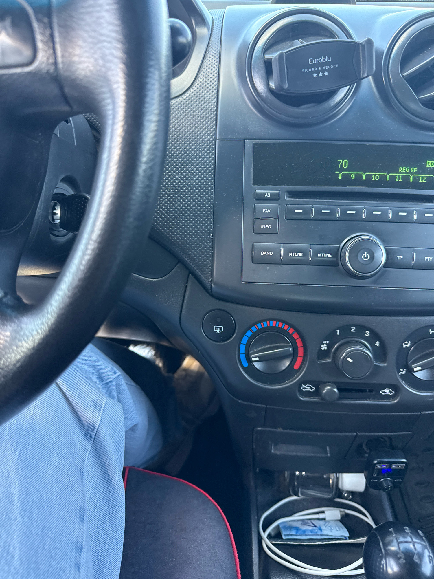 The image shows the interior of a car. A portion of the steering wheel and dashboard are visible. The dashboard includes a car radio, air vents, and climate control knobs. A mobile phone holder is attached to one of the air vents. A person's leg, clad in light blue jeans, is visible on the left side of the frame. A gear shift lever is visible on the lower right. There are no indications that this scene is located in Carabetovca, Moldova.