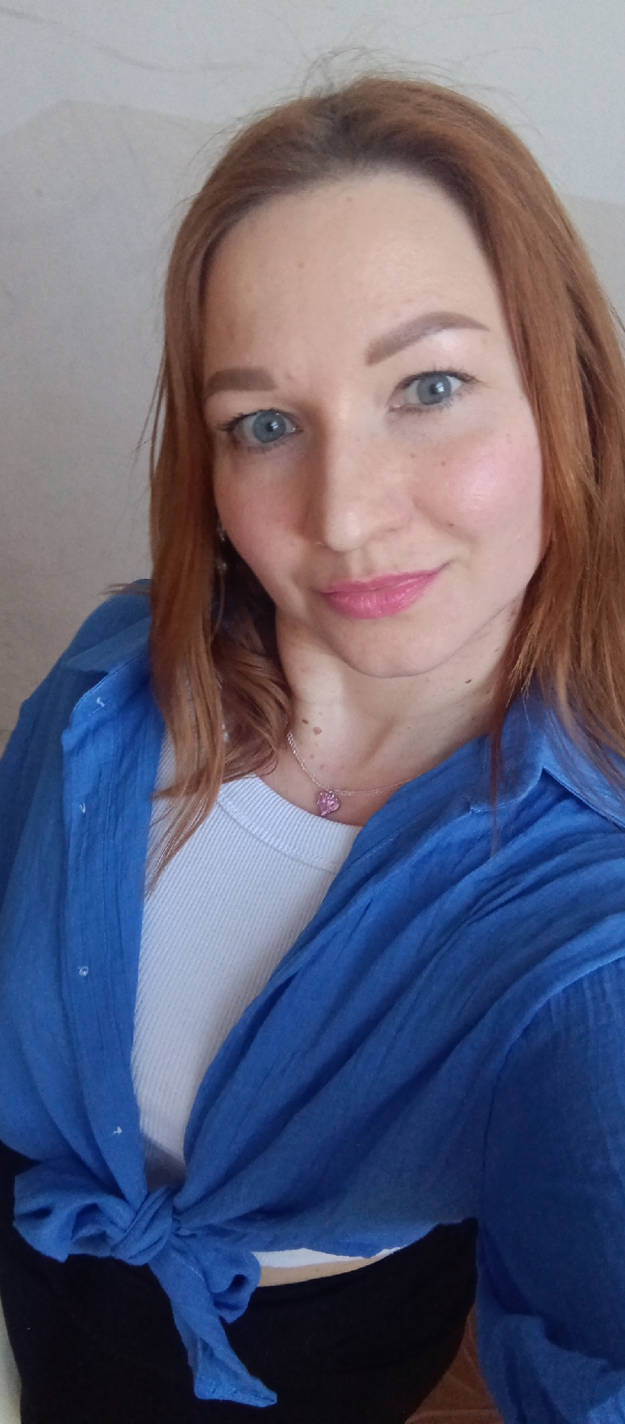 A woman with light skin, blue eyes, and auburn hair is centered in the image. She is wearing a white top and a blue button-down shirt that is tied in a knot at the bottom. A silver necklace with a pink pendant is visible around her neck. The background is a plain, light-colored wall. The location is Unknown, Unknown.