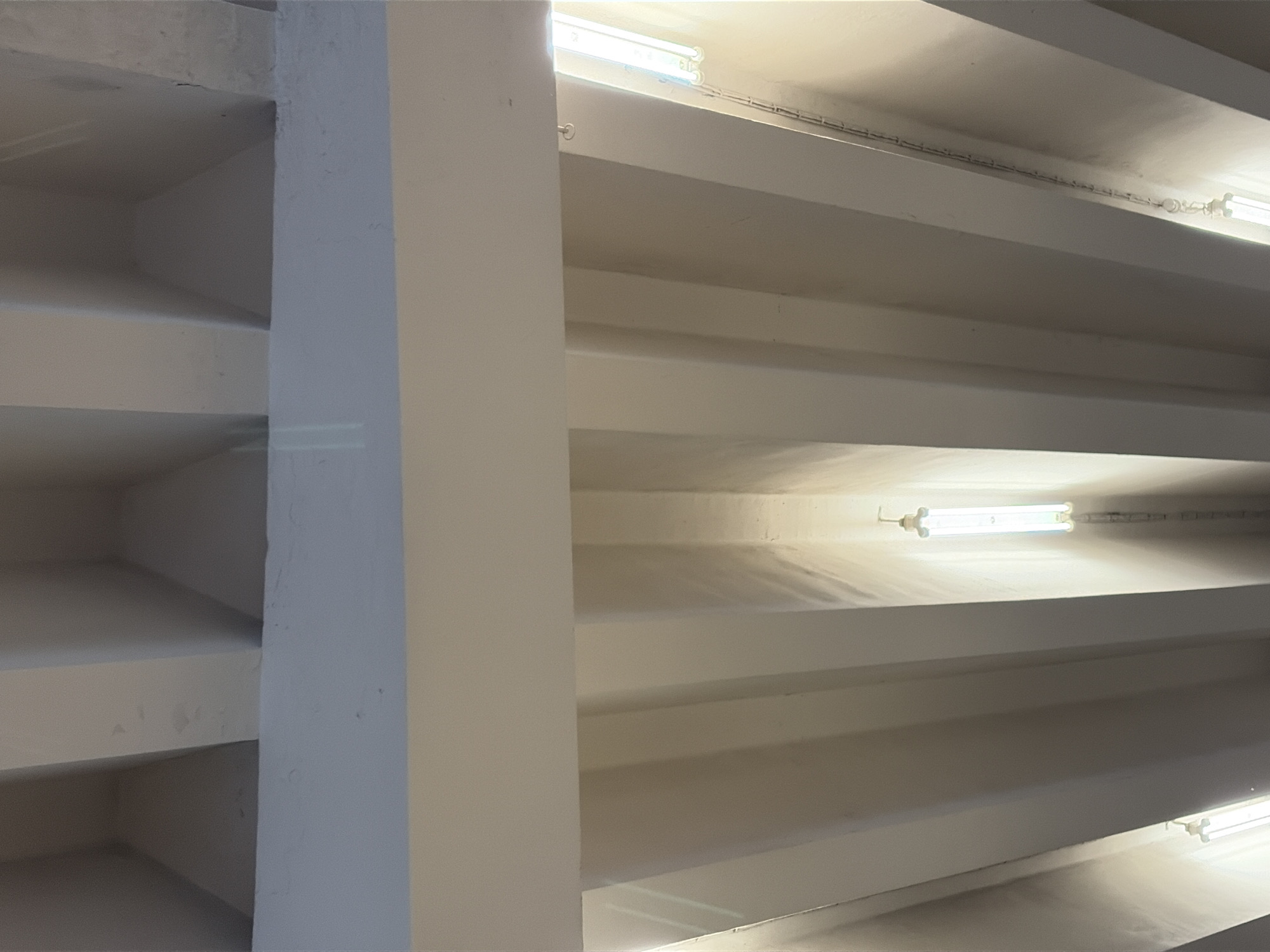 This is a low-angle, outdoor shot of the ceiling of a modern structure, possibly a walkway or a roof overhang, in Moscow, Russia. The ceiling consists of repeating, parallel white beams creating a series of recessed channels. Each channel contains a long, fluorescent light fixture, illuminating the space with a cool, white light. The texture of the beams appears smooth, with some minor imperfections visible. The lighting creates strong shadows within the channels, emphasizing the architectural structure. No people or other significant objects are visible in the frame, focusing solely on the architectural design and lighting.