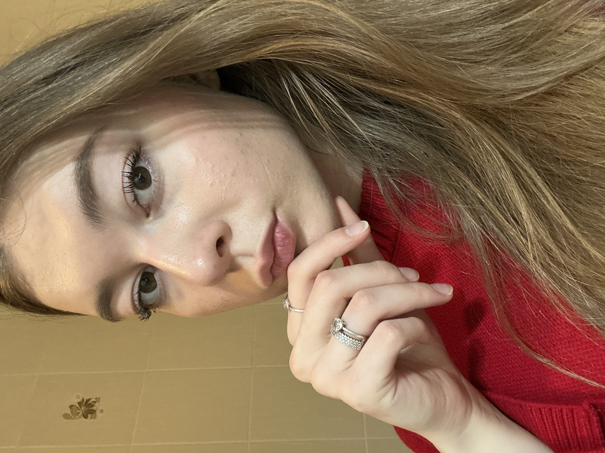 A close-up, rotated portrait features a young woman indoors. She has long, light brown hair that falls across her forehead and shoulder, framing her face. Her dark brown eyes are directed towards the camera, and her eyelashes appear enhanced with mascara. Her skin is fair, with some visible texture. Her lips are a soft pink and slightly parted.
She is wearing a red, knitted sweater with a visible texture. Her left hand is raised, with her fingers positioned near her chin and mouth. On the ring finger of her left hand, she wears two rings: one with a prominent clear stone and a textured band below it. A simple band ring is also visible on her middle finger. Her fingernails are natural and unpolished.
The background is a plain, light-colored wall, possibly tiled, with a subtle floral design visible on one tile in the lower part of the frame. The lighting suggests an indoor setting, likely with artificial light. Her expression is neutral to thoughtful. There is no visible text in the image. The location is Kazan, Russia.