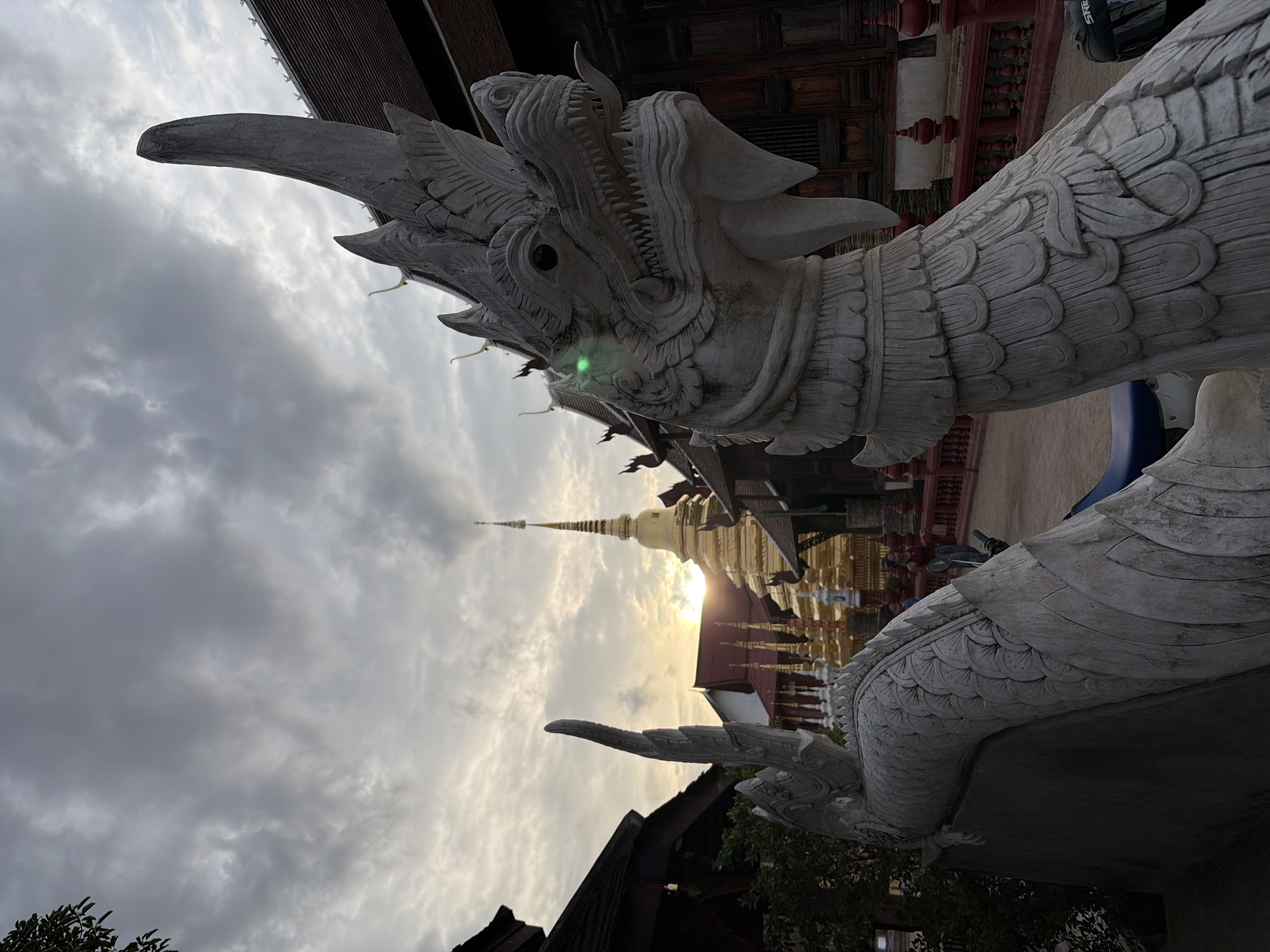 The image depicts a stone dragon sculpture in the foreground, with intricate scales and a fierce expression. Behind the dragon, a golden stupa gleams in the soft light of a cloudy sunset, likely in Chiang Mai City Municipality, Thailand. The stupa is surrounded by other traditional Thai temple structures, including wooden buildings with dark, tiered roofs and red railings. The sky is filled with dramatic, gray clouds, with a sliver of sunlight peeking through on the horizon. The overall atmosphere suggests a serene, spiritual setting at dusk. No people are clearly visible in the immediate vicinity of the sculpture.