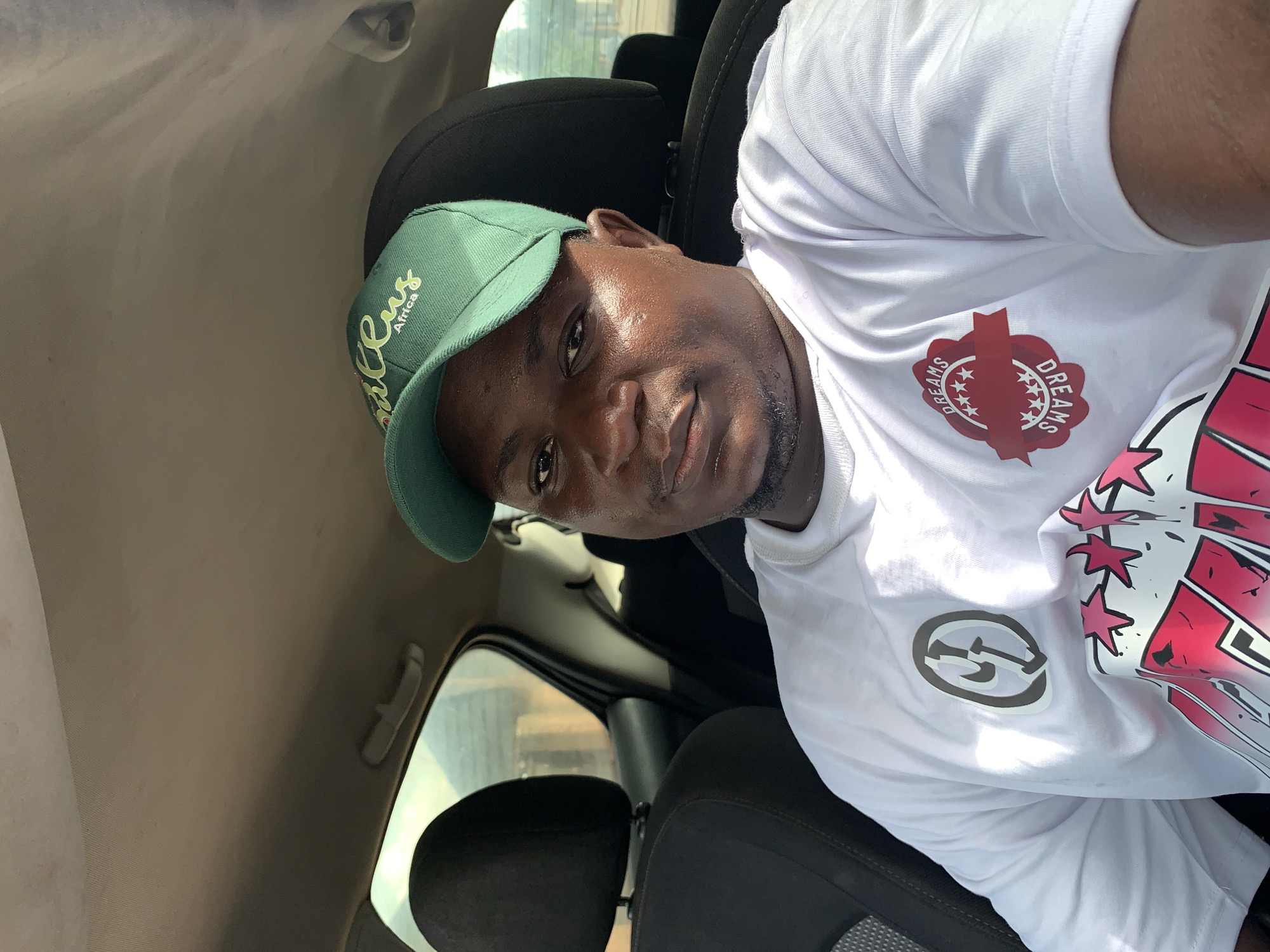 A portrait-style image, rotated counter-clockwise, depicts a man seated inside a car during the daytime in Akwanga, Nigeria. The man has a dark skin tone, short dark hair, and a short beard or stubble. He is wearing a light green baseball cap that features the text "Cactus Africa" in yellow script with "Africa" below in smaller white letters. He also wears a white, short-sleeved t-shirt adorned with multiple designs.
On the left chest of his t-shirt, there is a grey circular logo resembling a stylized "G" or two interlocking arrows. On the right chest area, a red emblem or shield shape is visible, displaying the word "DREAMS" twice on a curved banner, separated by stars. A larger, abstract design in red and pink with black outlines is partially visible on the lower right side of his t-shirt.
The man looks directly at the camera with a neutral to slightly smiling expression; his face shows a slight sheen, possibly from light reflection or sweat. The interior of the car is visible, including a black headrest and seat, and a light-colored car ceiling. A car window on the left reveals a blurred view of the outside environment. The bright lighting suggests it is daytime.