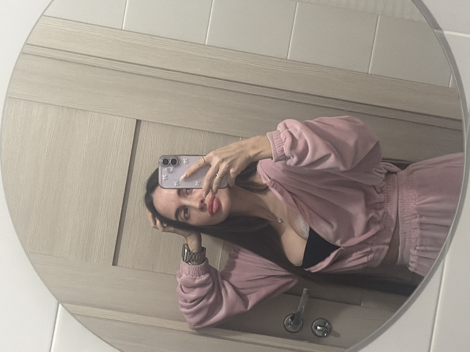 A young woman is taking a mirror selfie in a bathroom. The woman has dark hair and fair skin and is wearing a pink sweatsuit over a black top. She is holding a smartphone in her right hand. The bathroom has light wood paneling and a tiled wall. The location is not mentioned in the image.