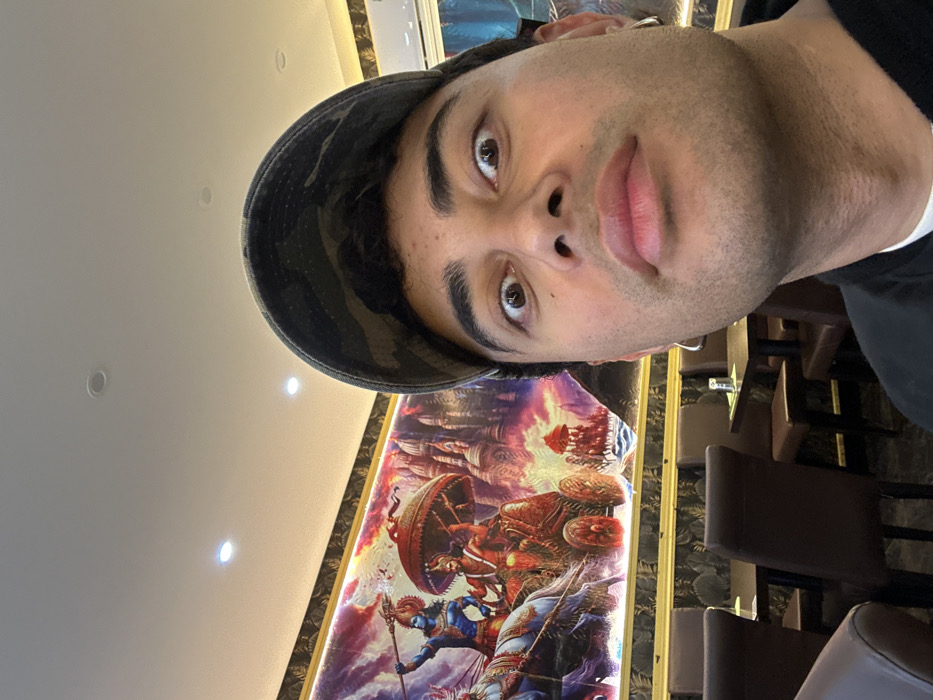 A male individual is depicted in a close-up selfie, occupying the right side of the frame. He has light brown skin, short dark hair visible beneath a dark camouflage-patterned baseball cap worn forward, and stubble on his face. His eyes are wide open, and his lips are slightly pursed, conveying an exaggerated expression. He wears a dark outer garment over a white undergarment and has a silver hoop earring in each ear.
The background reveals the interior of a commercial establishment, specifically a restaurant. A large, brightly lit mural extends across the far wall, framed by a gold border. The mural depicts a vibrant scene from ancient Indian mythology, featuring multiple figures, including two prominent individuals in chariots drawn by horses. One figure is blue-skinned and holds a spear, while the other, with reddish-brown skin, sits beneath a red parasol. Architectural elements resembling temples or palaces are visible in the background of the artwork. Below the mural, the wall is covered with a dark, patterned wallpaper. Multiple dark brown, high-backed chairs are arranged around dark-topped tables, some displaying small tabletop items. The ceiling is white with several circular recessed spotlights providing bright illumination.
The specific location within the city of Paris, France, cannot be determined from the visual information provided.