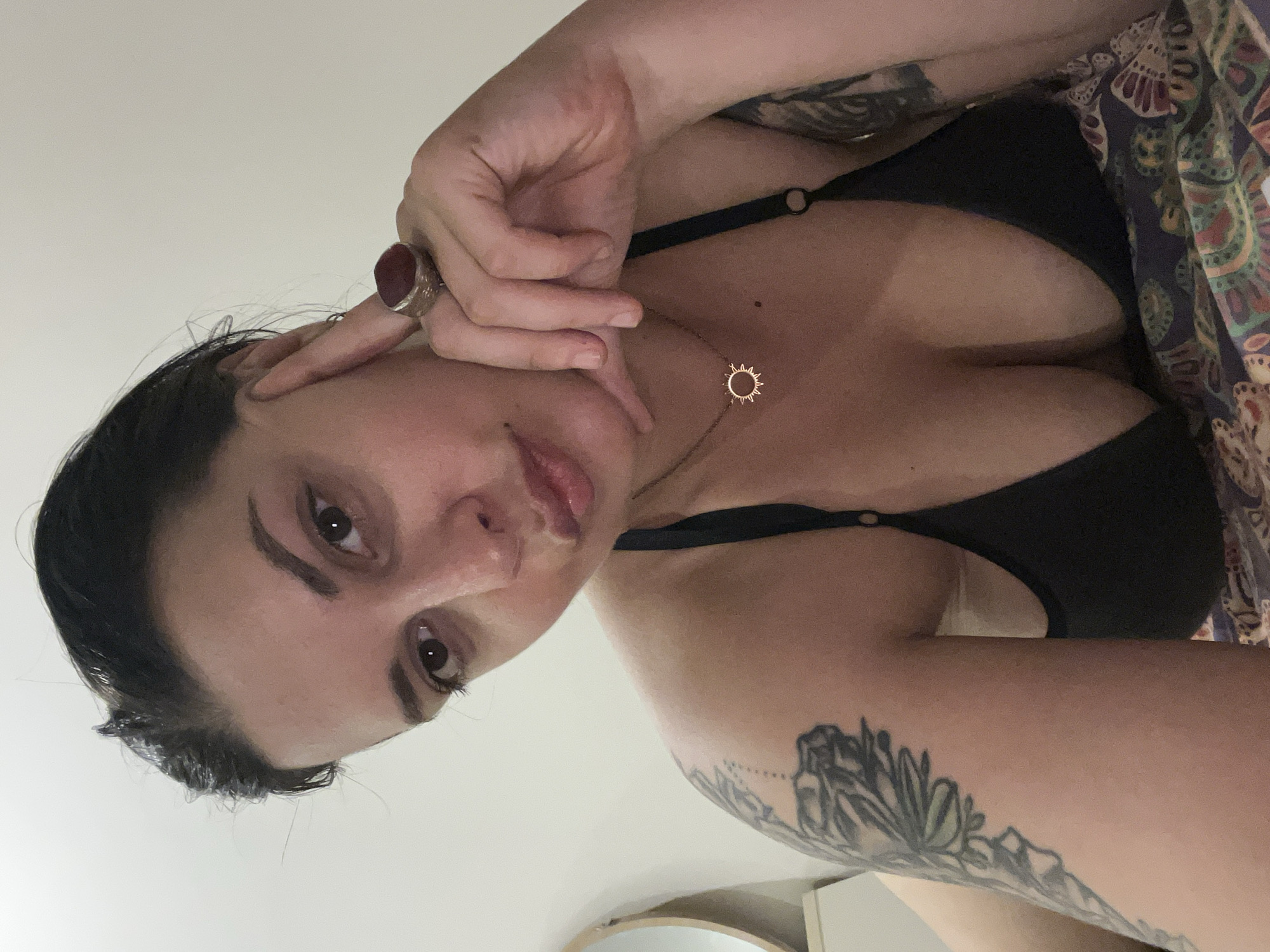 This is a close-up selfie of a woman with dark hair, resting her head on her hand. She is wearing a black bikini top and a necklace with a sun pendant. She has a large ring on her right index finger and tattoos on her left arm. The background is a plain, light-colored wall. There is a round mirror visible on the left side of the image, and part of a bed with a patterned comforter is visible at the bottom. The image does not contain any elements that suggest a specific location in the city of Đà Nẵng, Vietnam.