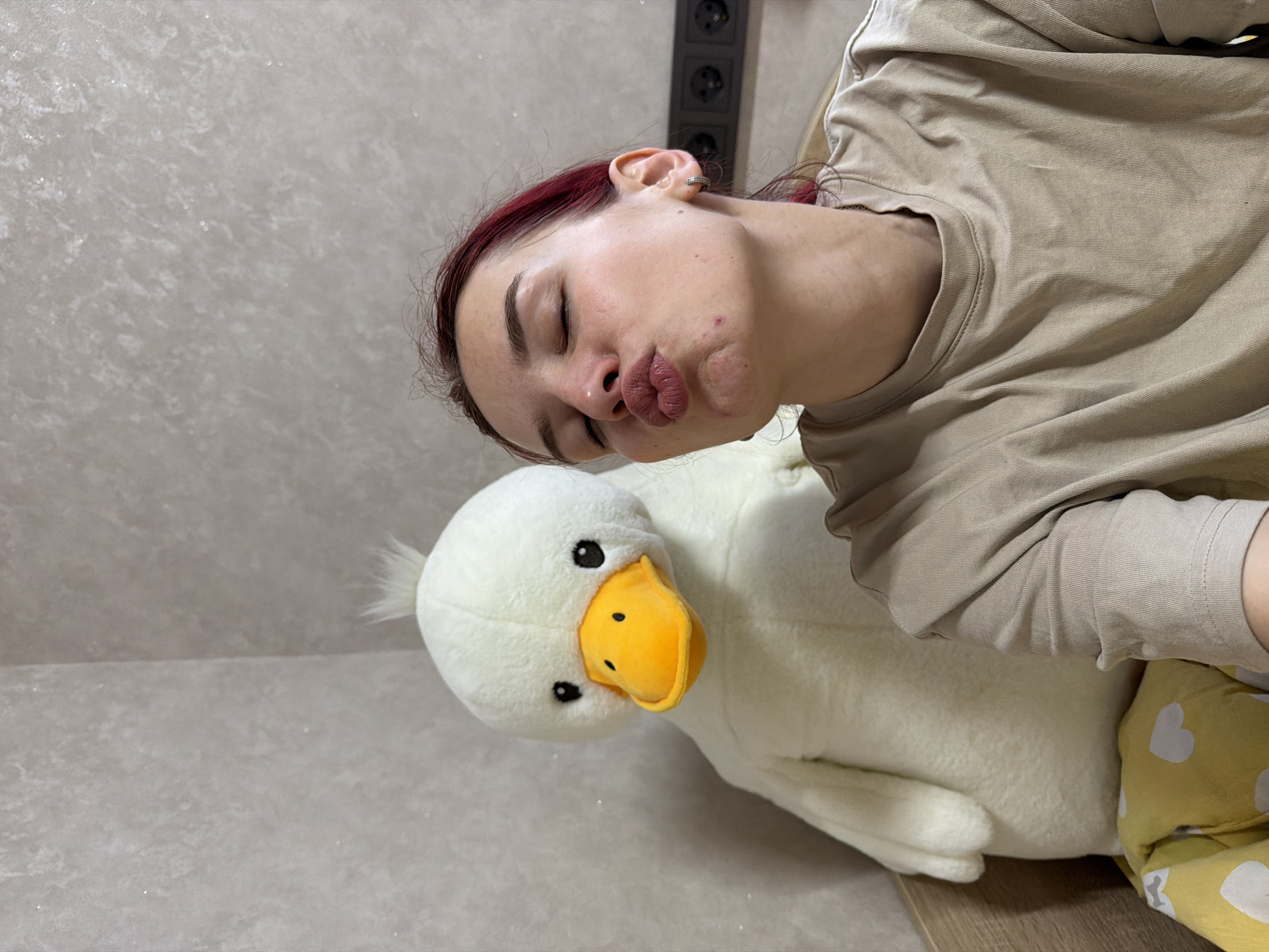 A woman with closed eyes and pursed lips is positioned at an angle, holding a plush white duck with a yellow beak. She is wearing a beige shirt and yellow pants with white hearts. In the background is a light-colored wall and an electrical outlet. There is no discernible information in the image to determine the exact location in Chaikovsky, Russia.