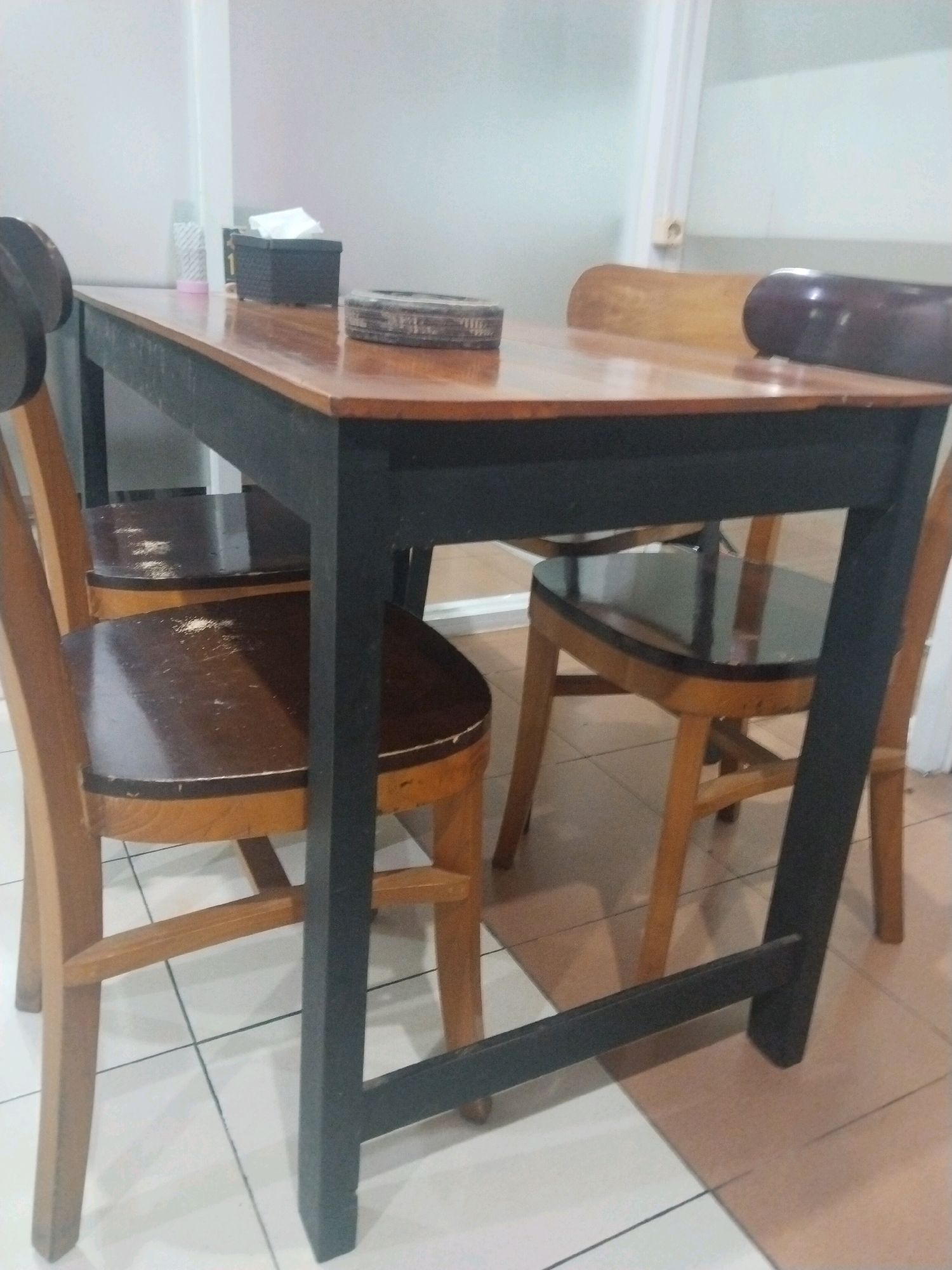A wooden dining table with black legs is set with two chairs on the left and two chairs on the right. The table has a wooden top and appears to be in use. A tissue box and a round object, possibly an ashtray, are placed on the table. The floor is tiled in a light color. This scene is located in Desa Ngesrep, Indonesia.