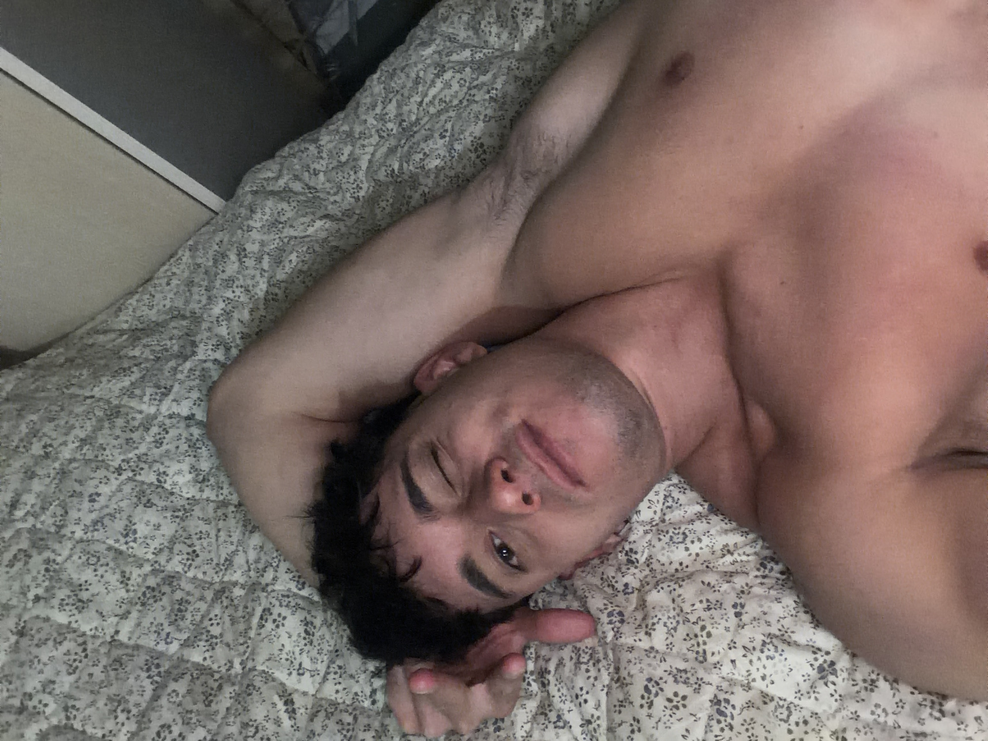 A shirtless man is lying on his back on a bed covered with a patterned bedspread. His left arm is raised above his head, and his right arm is bent with his hand near his face. The man is winking and looking at the camera. Part of a light-colored wall and a dark-colored object are visible in the upper left corner of the image. This image was not taken in Sofia, Bulgaria, and there are no indications of such in the image.