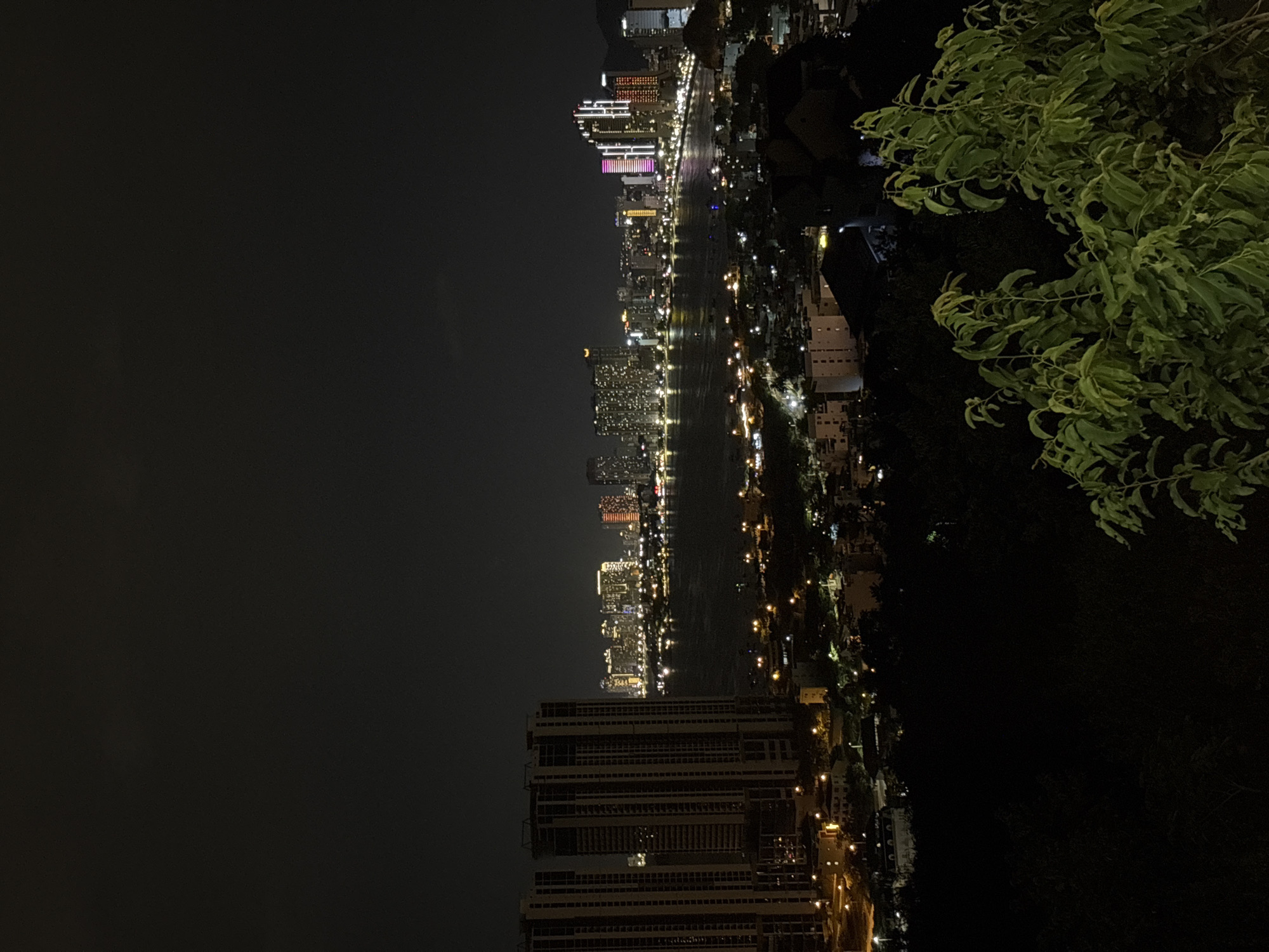 The image shows a nighttime urban scene in the city of Bac Nha Trang Ward, Vietnam. A body of water, likely a river or bay, runs vertically through the middle of the frame, reflecting the lights from buildings along the shoreline. The left side of the frame is predominantly dark, possibly indicating undeveloped land or open sky. A dense cluster of trees and foliage occupies the upper right portion of the frame. The urban landscape consists of high-rise buildings, residential structures, and streetlights. The lighting conditions suggest that the image was taken at night. The buildings closest to the bottom of the frame appear to be mostly apartments.