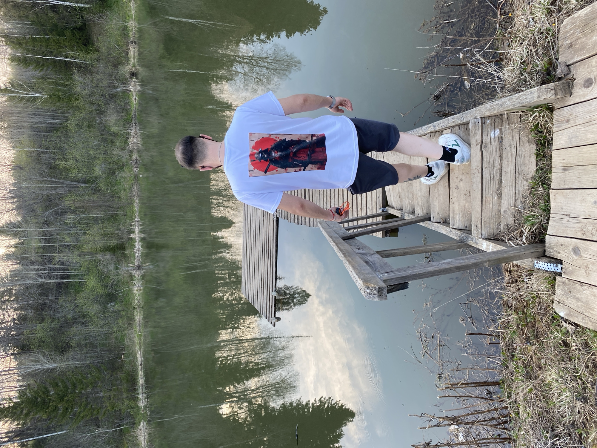 A man stands on a wooden dock extending into a body of water. He faces away from the camera, wearing a white T-shirt with a large graphic on the back, dark shorts, and white sneakers with black socks. The dock has a small wooden ladder leading down into the water. The water reflects the trees lining the opposite bank. The location is Баклуши, Russia.