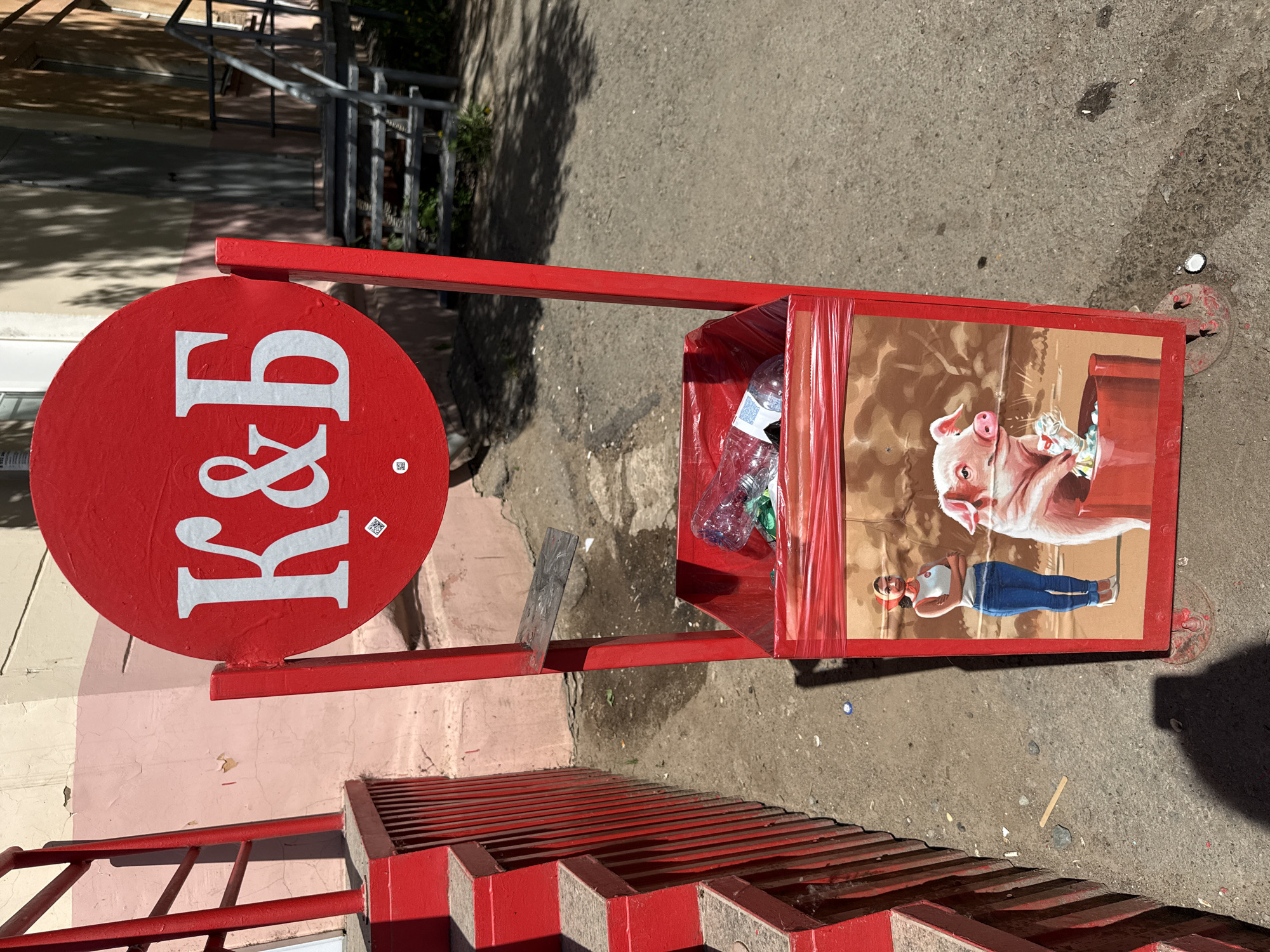 An outdoor street scene in Votkinsk, Russia, shows a red metal trash receptacle positioned on a concrete surface near a set of red metal stairs. The receptacle has a red circular sign with white letters "K&B" attached to it. The trash bin itself is decorated with an image of a pig holding a bottle and a woman with blue jeans lying down. The bin contains plastic bottles and other trash. The red stairs lead to a light-colored building with some discolored paint. The surrounding environment consists of asphalt and concrete.
