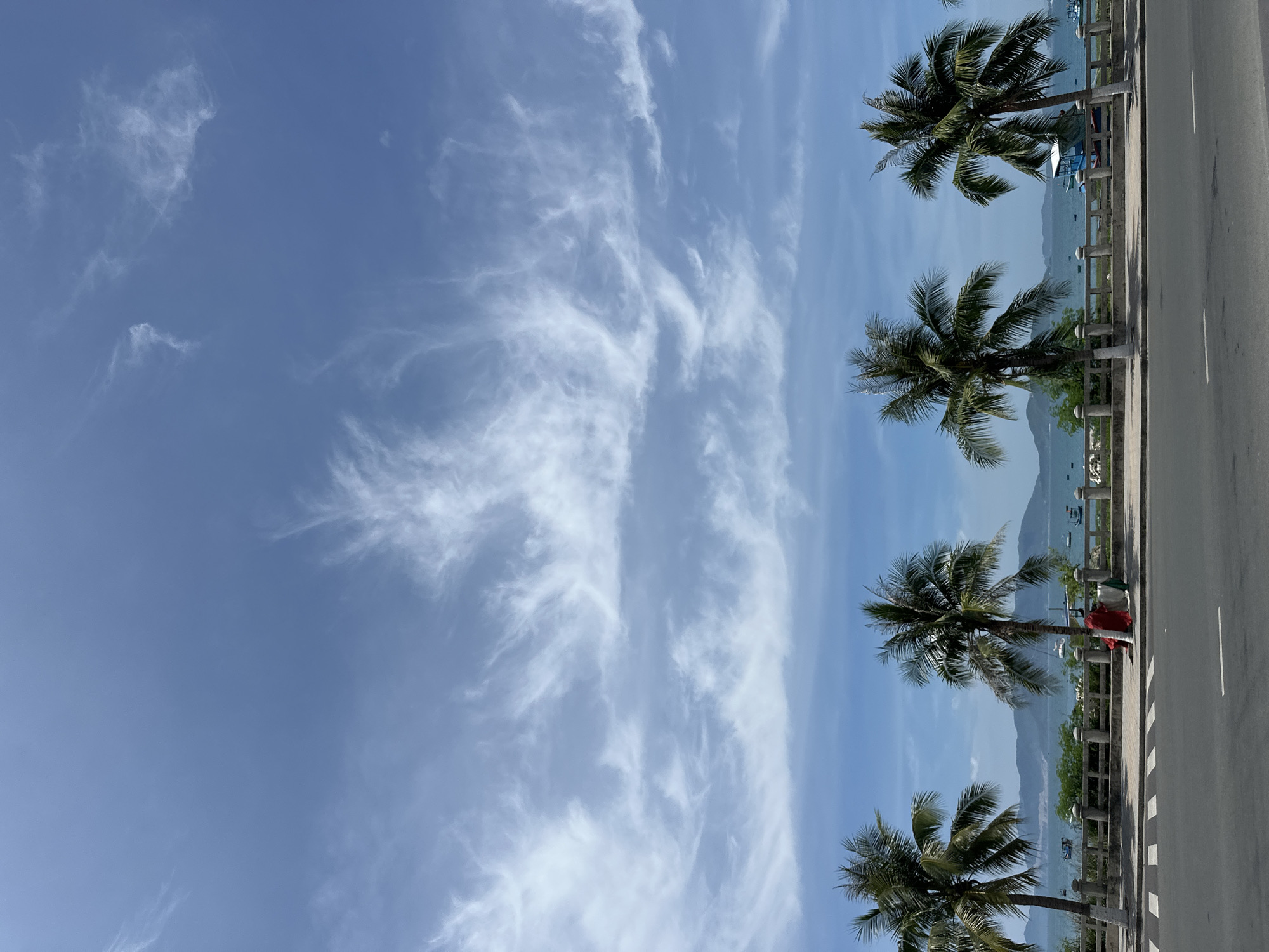 The image shows a tropical coastal scene in Bac Nha Trang Ward, Vietnam. In the foreground, a paved road runs horizontally across the bottom of the frame. Behind the road, a sidewalk is visible, bordered by a concrete railing with stone posts. Several palm trees line the edge of the sidewalk, their fronds swaying in the breeze. Beyond the railing, a body of water, likely the ocean, stretches out to the horizon. Several small boats are scattered across the water. In the distance, a chain of mountains is silhouetted against the sky. The sky itself is a vibrant blue, with wispy white clouds drifting across it. The overall atmosphere is one of a bright, sunny day in a coastal resort area.