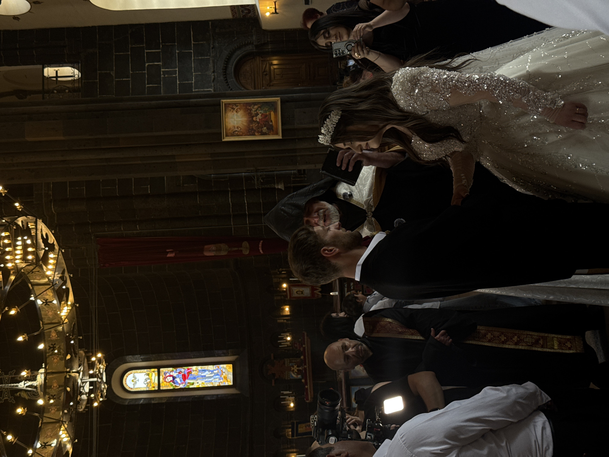 In the interior of a church in Gyumri, Armenia, a wedding ceremony is taking place. The groom, dressed in a black suit with a white shirt, stands facing the bride, who is wearing a sparkling white wedding dress and a tiara. A clergyman, in traditional dark robes with ornate trim, stands between them, holding a book and seemingly officiating the ceremony. Several guests are present, with some recording the event on their phones. The church features dark stone walls, a stained-glass window depicting a religious scene, a large chandelier, and religious artwork on the walls. The overall atmosphere appears solemn and celebratory.