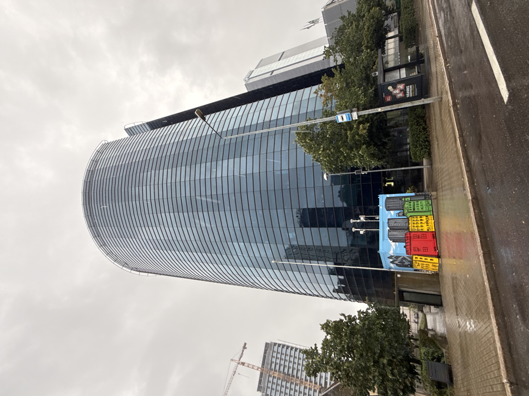 The image shows a panoramic view of a modern cityscape in Tallinn, Estonia, under an overcast sky. A prominent, curved glass-fronted skyscraper dominates the center, reflecting the surrounding environment. To its left, another taller building is under construction, identifiable by a yellow crane. In the foreground, a colorful mural depicting simplified urban buildings adorns a rectangular structure, possibly a utility box or container. The ground is paved with cobblestones and shows signs of recent rain, with puddles reflecting the muted light. Several trees with green foliage are scattered throughout the scene. A bus stop shelter with advertisements is visible on the right side of the image, alongside a lamppost. The overall atmosphere is somewhat somber due to the cloudy weather.