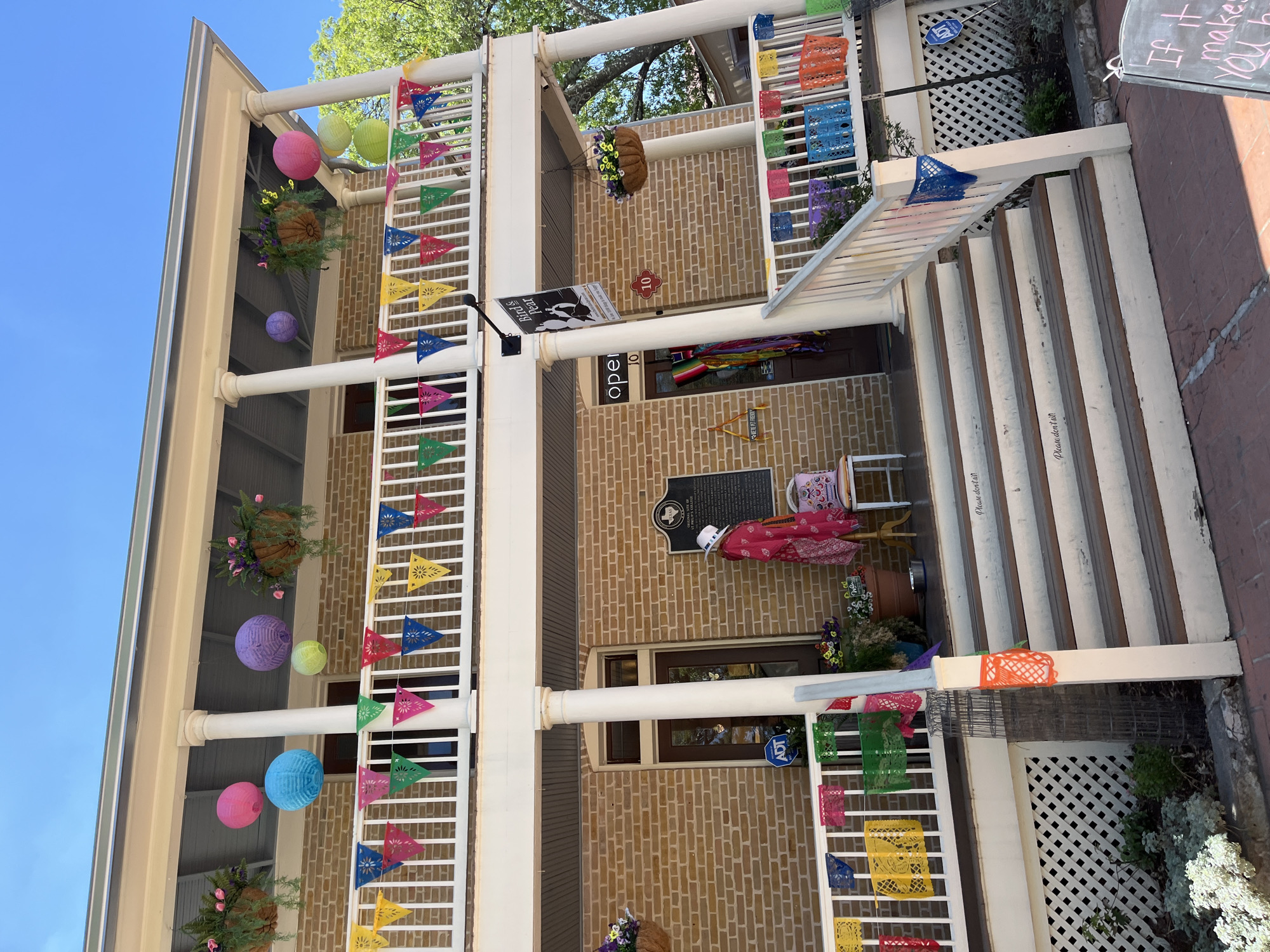 The image shows the exterior of a two-story brick building, likely a business in San Antonio, Texas, based on its apparent festive decor and architectural style. The building has white columns and railings, decorated with colorful "papel picado" banners and hanging paper lanterns, suggesting a Mexican or Hispanic-themed celebration or ongoing ambiance. A sign reading "Birds Pear" and "Open" hangs above the entrance, indicating a shop or restaurant name. Additional decorative elements include hanging flower baskets and a mannequin in a colorful dress. A Texas historical marker is visible on the building's façade. To the right, a blackboard displays a handwritten phrase, "If it makes YOU happy," adding a personal touch. Steps lead up to the entrance, and the environment is urban.