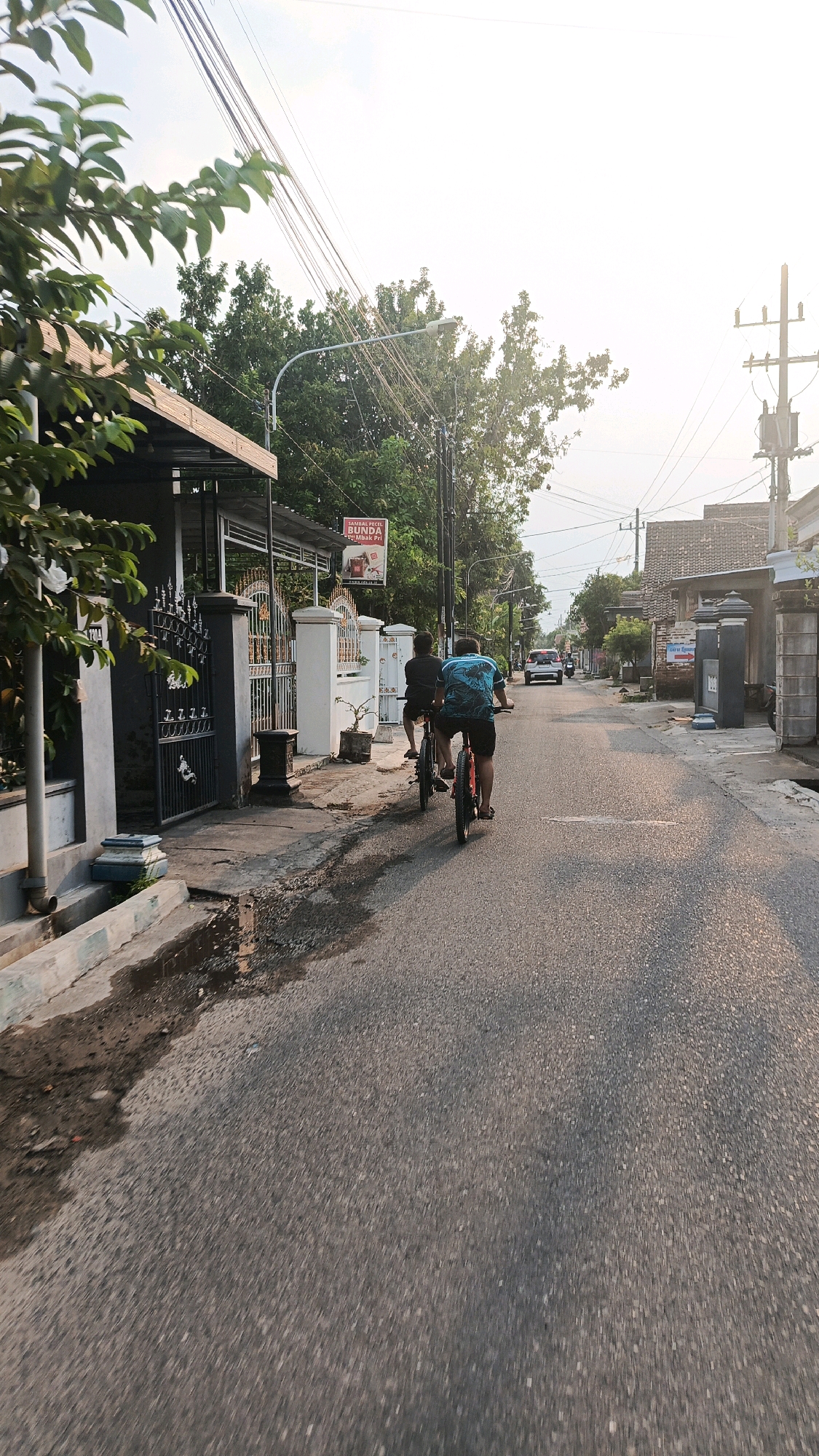Two individuals are cycling on an asphalt road in a residential area of Mangunsuman, Indonesia. The person in front is wearing a blue t-shirt with a graphic design and black shorts. The person behind is wearing a black t-shirt and dark shorts. Both are facing away from the camera. A white car is visible in the distance on the road. The environment includes houses with tiled roofs, trees, power lines, and streetlights. The road has some puddles on the left side, suggesting recent rain. The lighting indicates it is either early morning or late afternoon, with sunlight casting shadows. A sign advertising "Sambal Pecel Bunda" is mounted on a pole on the left side of the street, near a gate with intricate metalwork.