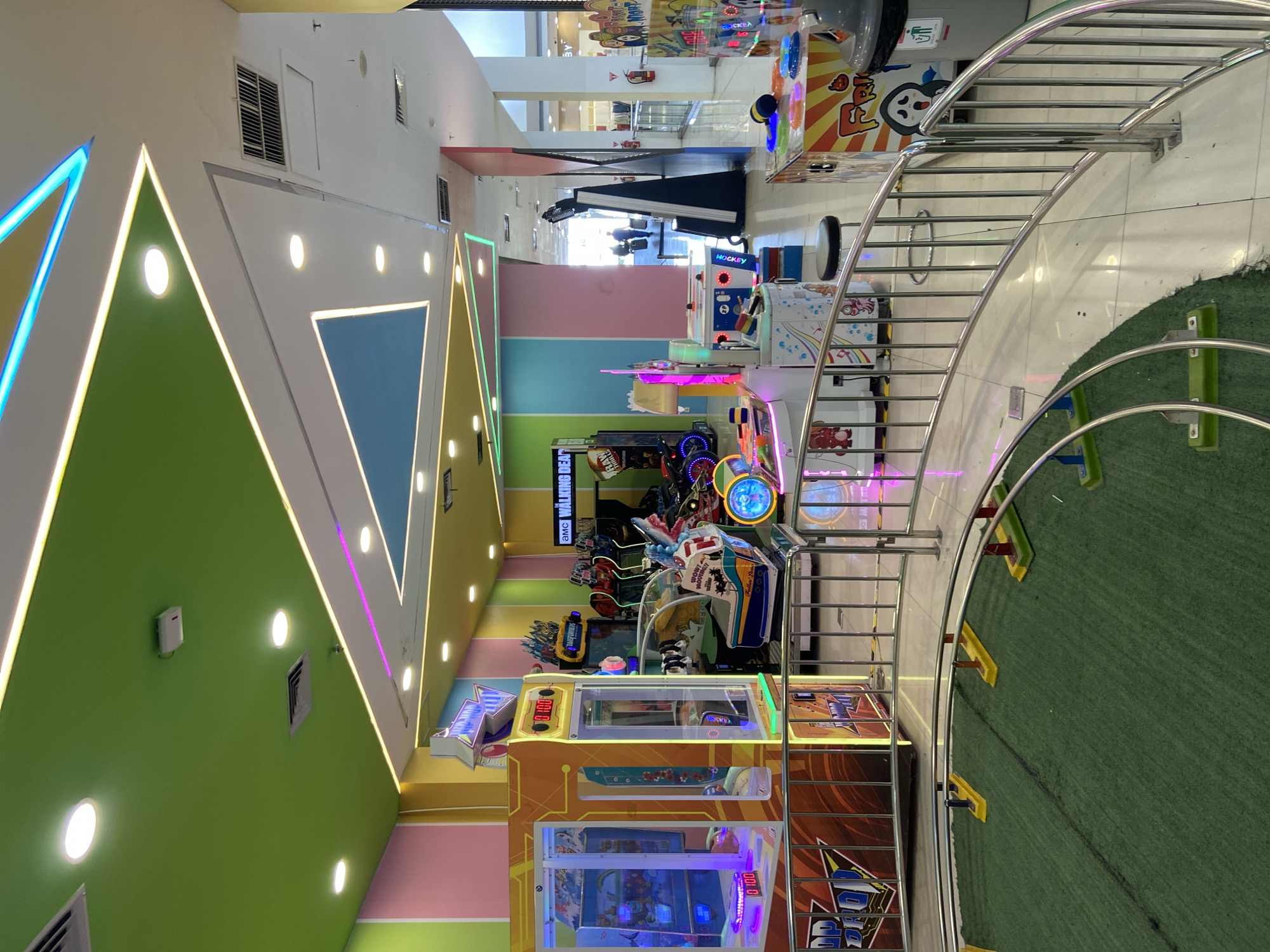 The image shows an indoor arcade in Kudus, Indonesia. The arcade features a variety of colorful game machines, including claw machines, racing games, and air hockey tables. The ceiling is painted in vibrant colors with neon lighting, creating a lively and energetic atmosphere. In the foreground, a small train track with green turf is visible, suggesting a children's ride. A metal railing encloses the track and separates it from the rest of the arcade. Several game machines are positioned behind the railing, and some people can be seen in the background, interacting with the machines or walking through the arcade. The overall impression is of a fun and engaging entertainment venue.