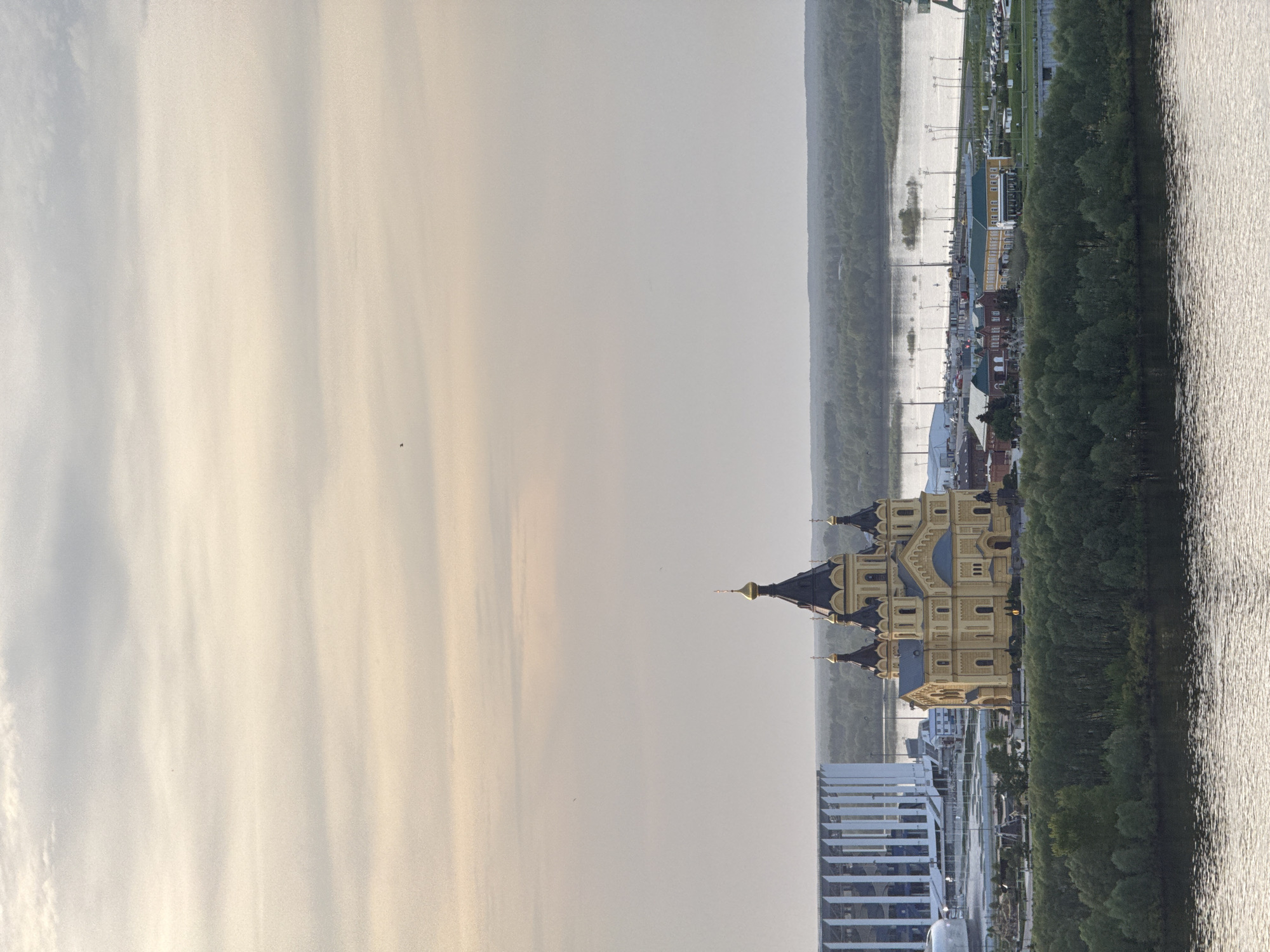 The image depicts the Alexander Nevsky Cathedral in Nizhny Novgorod, Russia, as viewed from a high vantage point. The cathedral, with its golden domes and detailed facade, stands prominently against a backdrop of modern buildings and trees. The Oka river runs alongside the cathedral. The sky above is light.