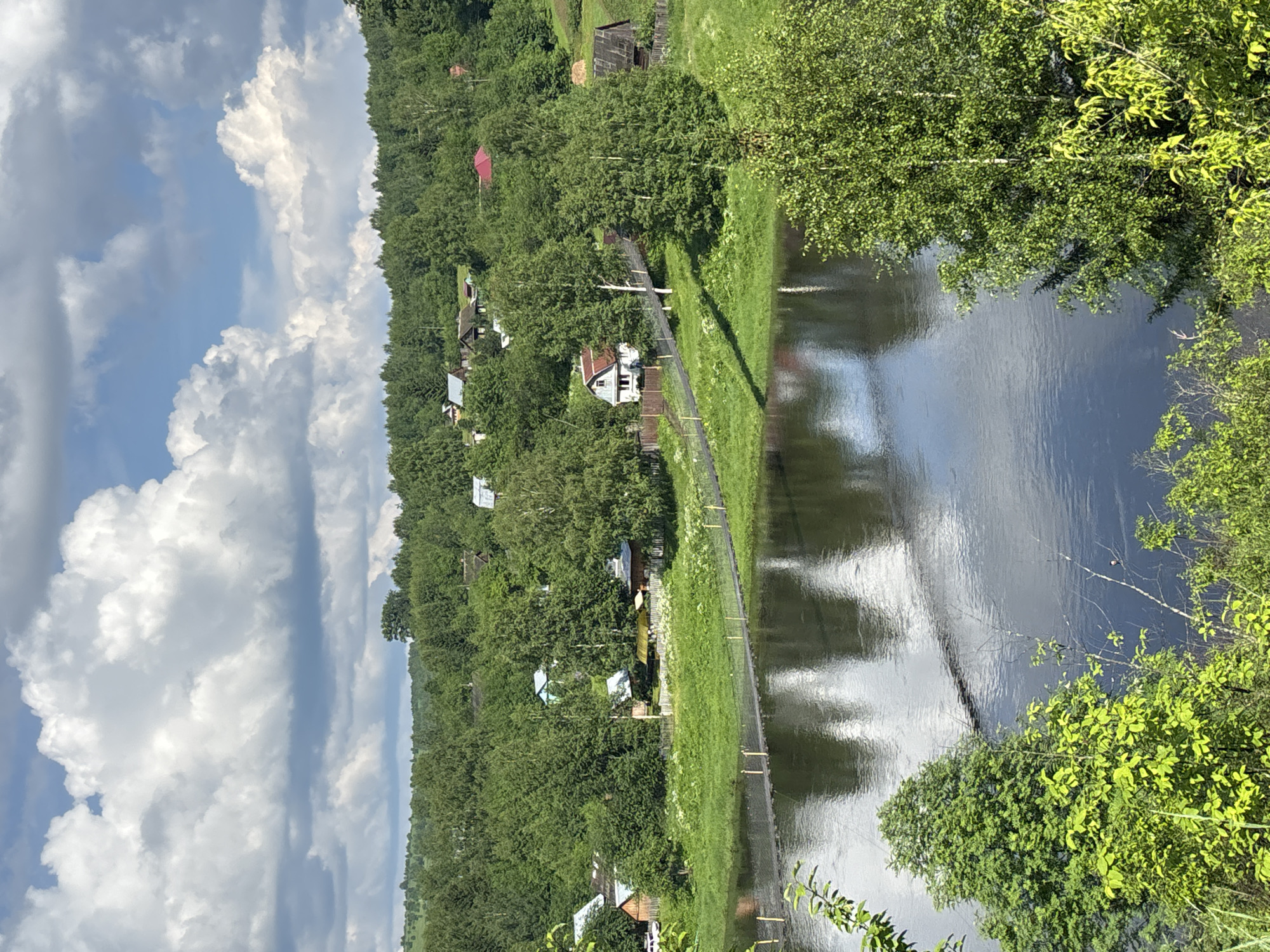 The image depicts a serene riverside landscape in Gubakha, Russia. A suspension bridge, constructed with dark planks and supported by cables, spans a calm, dark river. The river's surface reflects the overcast sky and the surrounding greenery. On the opposite bank, a verdant hill is densely covered with lush green trees, interspersed with several small, traditional wooden houses, some with red or grey roofs, and a few scattered structures like sheds or barns. The architecture suggests a rural or semi-rural setting. The foreground features vibrant green foliage and trees, framing the view of the river and the village. The sky above is a mix of blue and large, white cumulus clouds, indicating fair weather. The scene conveys a sense of tranquility and natural beauty.