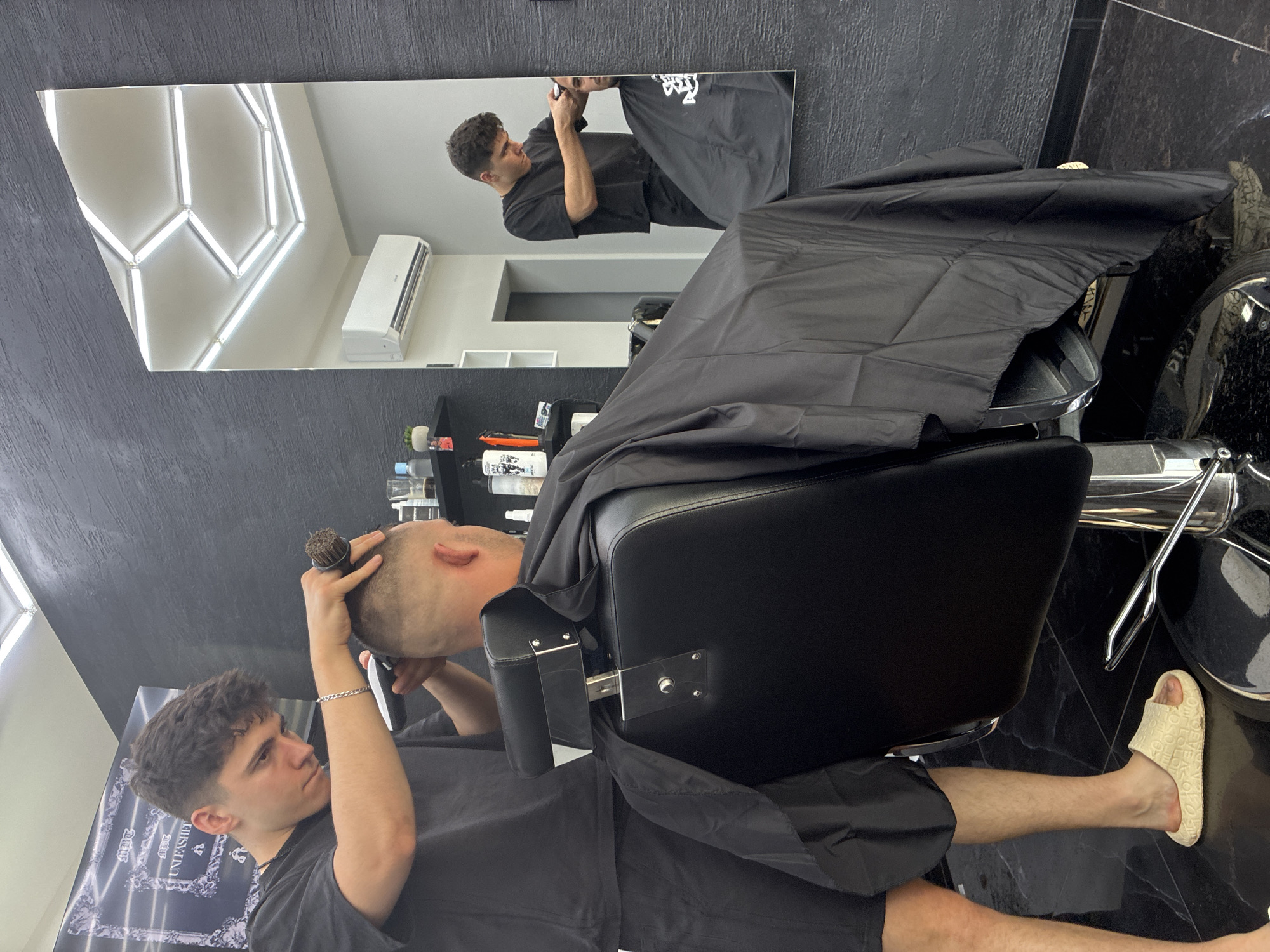 This indoor photograph, taken in Simitli, Bulgaria, captures a barbershop scene with a modern, dark aesthetic. The image is oriented sideways, with the main subjects appearing rotated.
In the foreground, a young male barber with dark, curly hair, wearing a black t-shirt and shorts, is intently working on a client's hair. He wears a silver chain bracelet on his left wrist and holds a hair brush near the client's head. The client, positioned in a black upholstered barber chair and draped with a black cape, has very short, buzzed hair and is looking upwards. His light-colored, slip-on sandals, adorned with a repeating text pattern, and lower legs are visible beneath the cape on the dark, possibly tiled floor.
A large rectangular mirror dominates the background, reflecting the barber from a different angle as he works. The reflection also shows parts of the barbershop's ceiling, featuring a striking geometric pattern of bright, hexagonal light fixtures, and a white air conditioning unit mounted on the wall. Behind the barber chair, shelves stocked with various hair care products are partially visible. A poster with the word "UNLEASHED" and an ornate design is also faintly visible in the reflection.
The atmosphere appears focused and professional, with both individuals engaged in the haircutting process. The bright artificial lighting suggests either daytime or a well-lit interior. The predominant colors are black, white, and grey, contributing to the contemporary feel of the establishment.