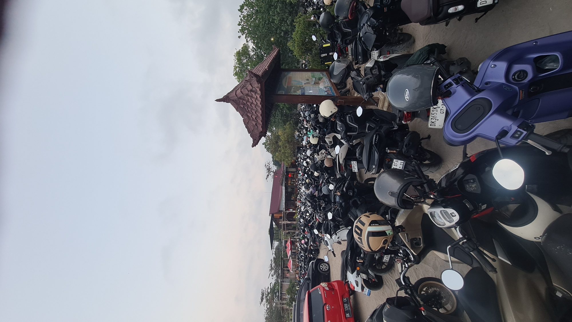 The image shows a vast outdoor parking lot filled with hundreds of motorcycles and scooters, predominantly black and white, with a few colored ones. Several helmets are placed on the handlebars or seats of the parked vehicles. In the background, there are buildings with red roofs and some greenery, indicating an urban or semi-urban environment. A signpost with a wooden roof and a display board is visible to the right. The sky is overcast. The license plates of some vehicles are partially visible, suggesting the location is Denpasar, Indonesia. For example, a red car has a license plate starting with "DK," a common prefix for vehicles registered in Bali, Indonesia.