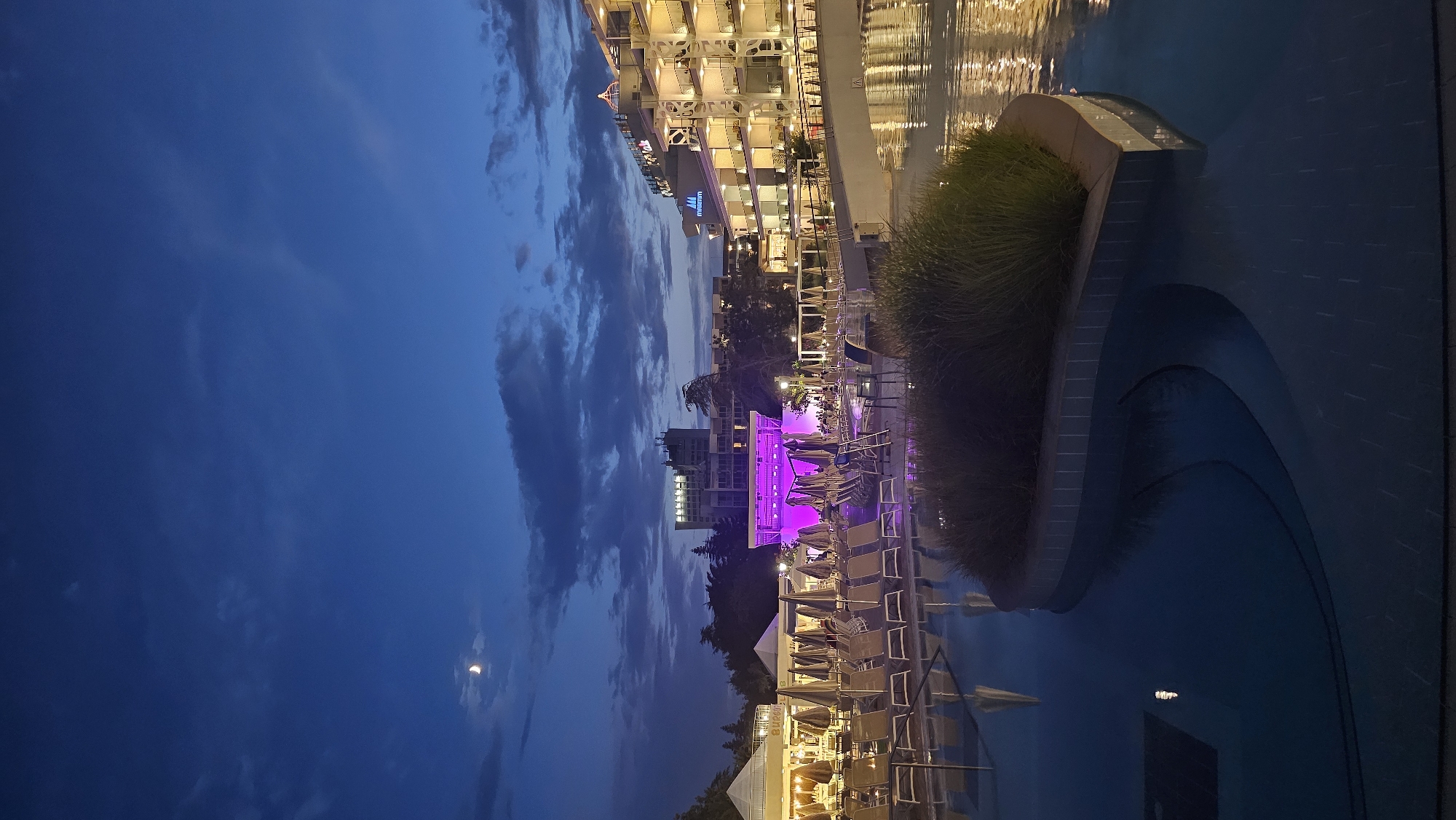 The image captures a twilight scene at the Grand Hotel Mimosa in Obrochishte, Bulgaria. In the foreground, a swimming pool reflects the ambient light from the surrounding buildings and sky. The pool features curved edges and an island planted with tall grass. Beyond the pool, rows of lounge chairs and umbrellas are neatly arranged, suggesting a resort setting. In the mid-ground, a structure with purple lighting is visible, possibly an outdoor stage or entertainment area. To the right, a multi-story hotel building is illuminated, with balconies and windows glowing warmly. The sky above is a deep blue, with a sliver of the moon visible and scattered clouds. The overall atmosphere is serene and inviting, indicative of a hotel resort evening.