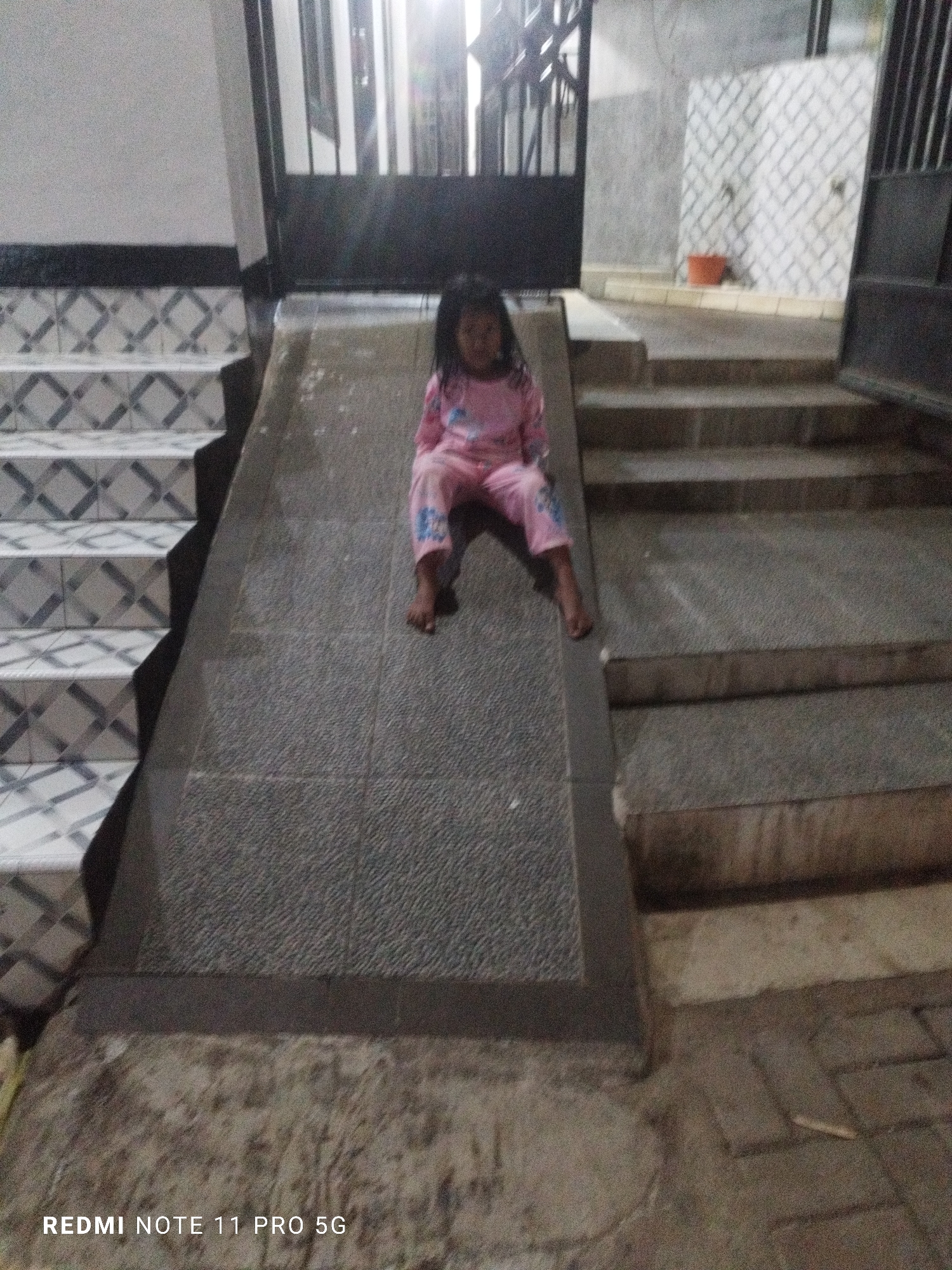 A young girl in pink pajamas sits on a grey ramp in front of a staircase in Kaduhejo, Indonesia. The staircase has white tiles with a grey diamond pattern. A black metal gate is visible behind the girl. The ramp is made of speckled grey concrete. The ground in front of the ramp appears to be paved with brown bricks. The overall scene is outdoors with artificial lighting present.