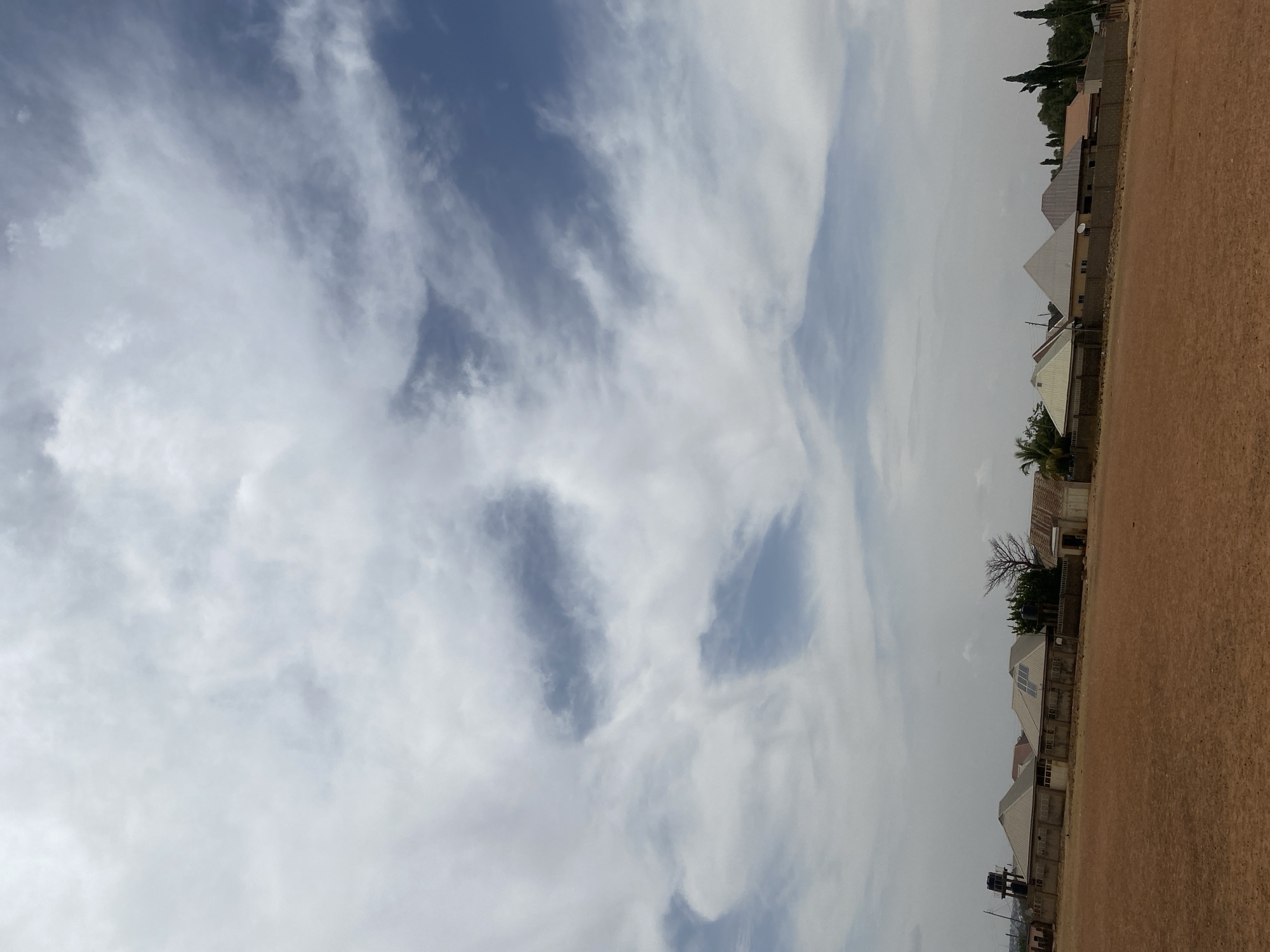 The image shows a view of the sky and part of a built environment in Rido, Nigeria. The sky is predominantly cloudy with patches of blue visible. Below the sky, a line of buildings with varying roof styles can be seen. Adjacent to these buildings is a relatively flat, brown-colored area, possibly a field or bare ground. No people are visible in the image.