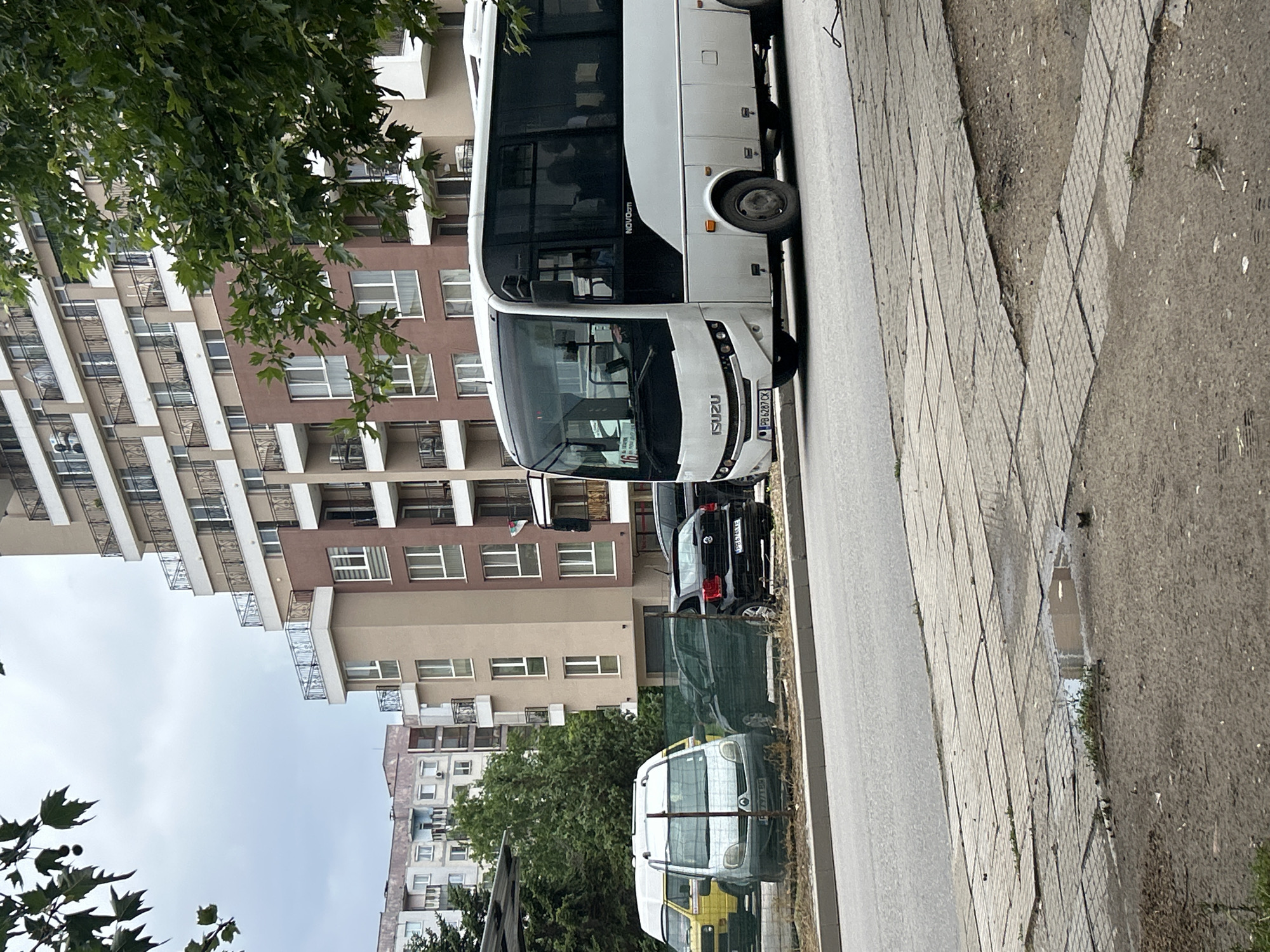 The image captures an urban street scene under an overcast sky, likely in Plovdiv, Bulgaria, as indicated by visible license plates. A white Isuzu Novociti Life bus is the central subject, parked on the right side of an asphalt road. The bus displays "ISUZU" on its front grille and "NOVOCiti" on its side, along with the Bulgarian license plate "PB 6287 CX." Behind the bus, a black car is partially visible, also parked. Further down the street, behind a green wire fence, a white and yellow van or small truck is parked, with a partially visible license plate "E 7971 CA."
To the right of the road, a grey paved sidewalk features rectangular tiles. Beyond the sidewalk, an unpaved strip of dirt and gravel contains several puddles, suggesting recent rain. In the background, large multi-story residential buildings with multiple balconies are visible. One prominent building features contrasting brown and beige sections. Green foliage from trees frames the scene, with leaves in the foreground and branches obscuring parts of the buildings. The time of day appears to be daytime, with soft, diffused light consistent with an overcast sky. No human activity or emotion is discernible; the scene portrays a quiet, parked street.