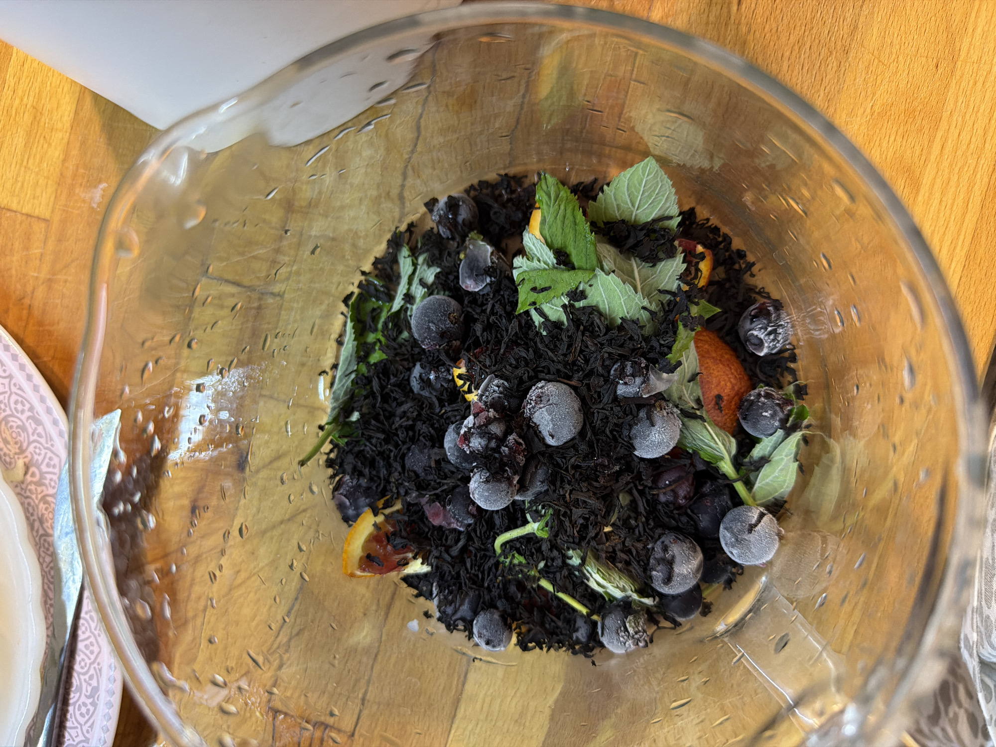 A close-up, overhead shot shows a glass pitcher filled with loose black tea, frozen blueberries, mint leaves, and small pieces of dried orange. The pitcher is sitting on a wooden surface. The pitcher has condensation on the outside. In the upper left corner, a portion of a decorative white plate with pink and gray patterns is visible. The scene is well-lit, and the focus is on the contents of the pitcher.