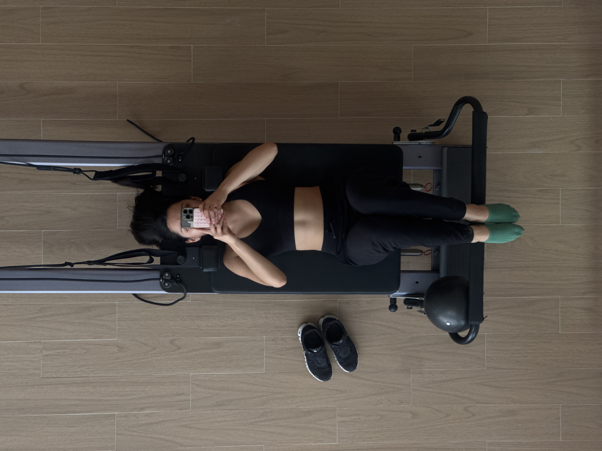 A top-down shot shows a woman lying on a Pilates reformer machine, indoors on a wood-look tiled floor. The woman is wearing a black sports bra, black pants, and green socks. She is holding a pink cell phone up to her face. Her black hair is spread out on the black reformer carriage. A pair of black and white sneakers sit on the floor near the foot end of the machine. The machine itself is mostly black and gray. The location is unknown and not identifiable from the image. There is no indication that this image was taken in San Bernardino Tlaxcalancingo, Mexico.