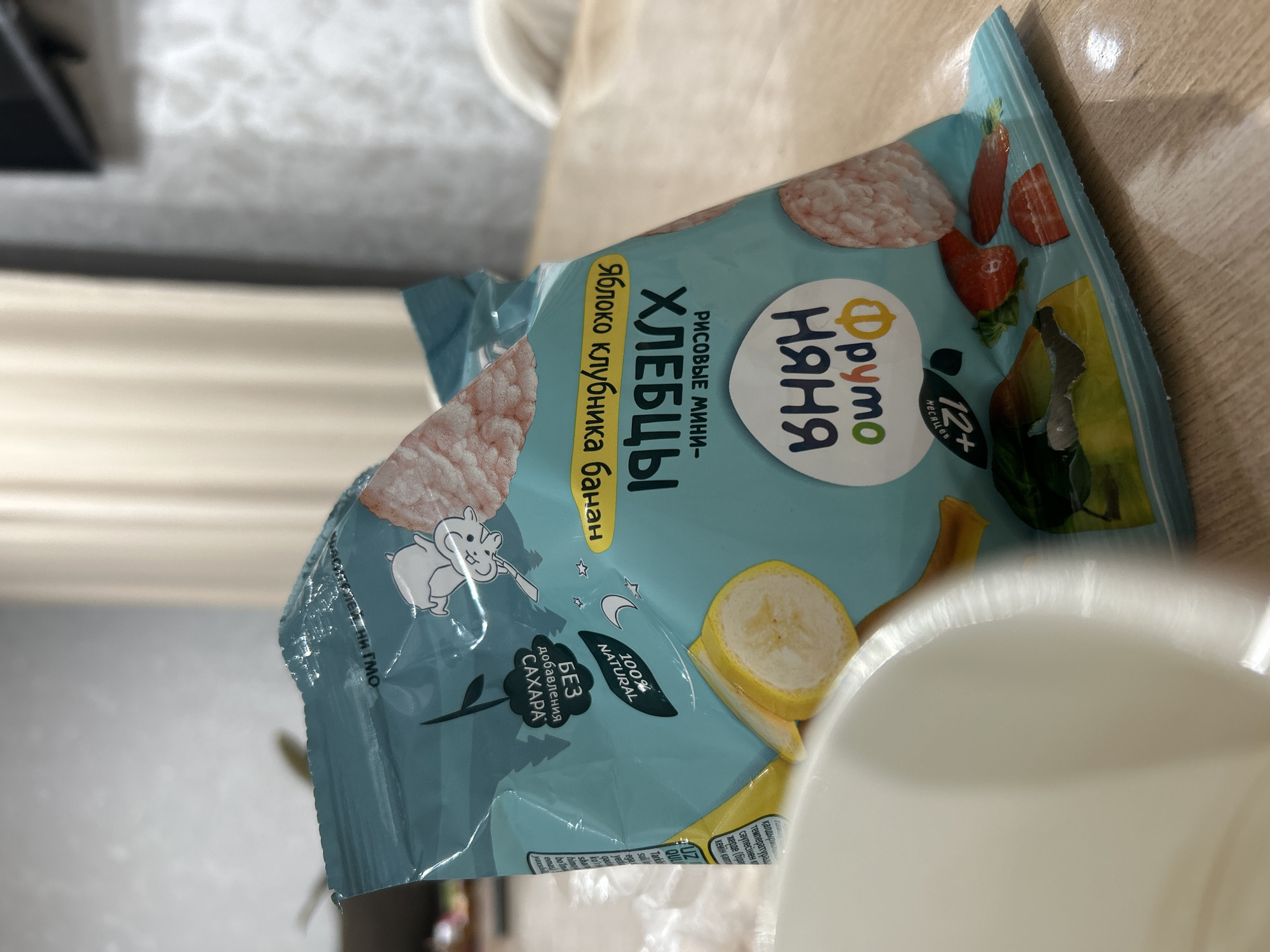 An indoor, slightly tilted shot taken in Chaikovsky, Russia, features a light blue, flexible package of "Фруто Няня" (Fruto Nyanya) brand rice mini-crisps resting on a light-colored wooden table. The package is adorned with illustrations of rice crisps, strawberries, and banana slices, along with a cartoon squirrel-like character.
Key text on the packaging, written in Cyrillic, identifies the product as "РИСОВЫЕ МИНИ- ХЛЕБЦЫ" (RICE MINI-CRISPS) with flavors "Яблоко клубника банан" (Apple strawberry banana). It indicates suitability for "12+ Месяцев" (12+ Months) and highlights that it is "БЕЗ добавления САХАРА" (WITHOUT added SUGAR) and "100% NATURAL". Partially visible text also includes "ей, ни ГМО" (likely indicating GMO-free).
A white cup or bowl is partially visible in the foreground, bottom right. The background is blurred, showing a light-colored wall or curtain. The package shows signs of being slightly crumpled, and there is a bright glare reflecting off its surface.