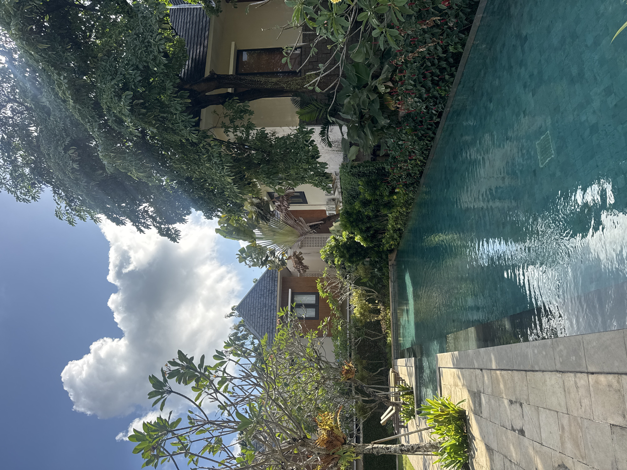 The image depicts an outdoor swimming pool area in Kuta Selatan, Indonesia. The pool is rectangular and filled with turquoise water, tiled on the bottom, with visible steps leading into it. The pool is surrounded by a stone-tiled pathway on the left, bordered by lush green foliage and some tropical plants. To the right of the pool, a dense wall of green plants with red flowers runs along its edge.
Behind the pool and pathway, there are buildings with tiled roofs and wooden accents. The architecture suggests a resort or villa setting. Large trees with full canopies are prominent in the scene, with one particularly large tree casting shadows over a portion of the property. The sky above is bright blue with fluffy white clouds. Sunlight is filtering through the trees, creating dappled light effects on the ground and water. There are no people visible in the image. The overall impression is one of a serene and tropical environment.