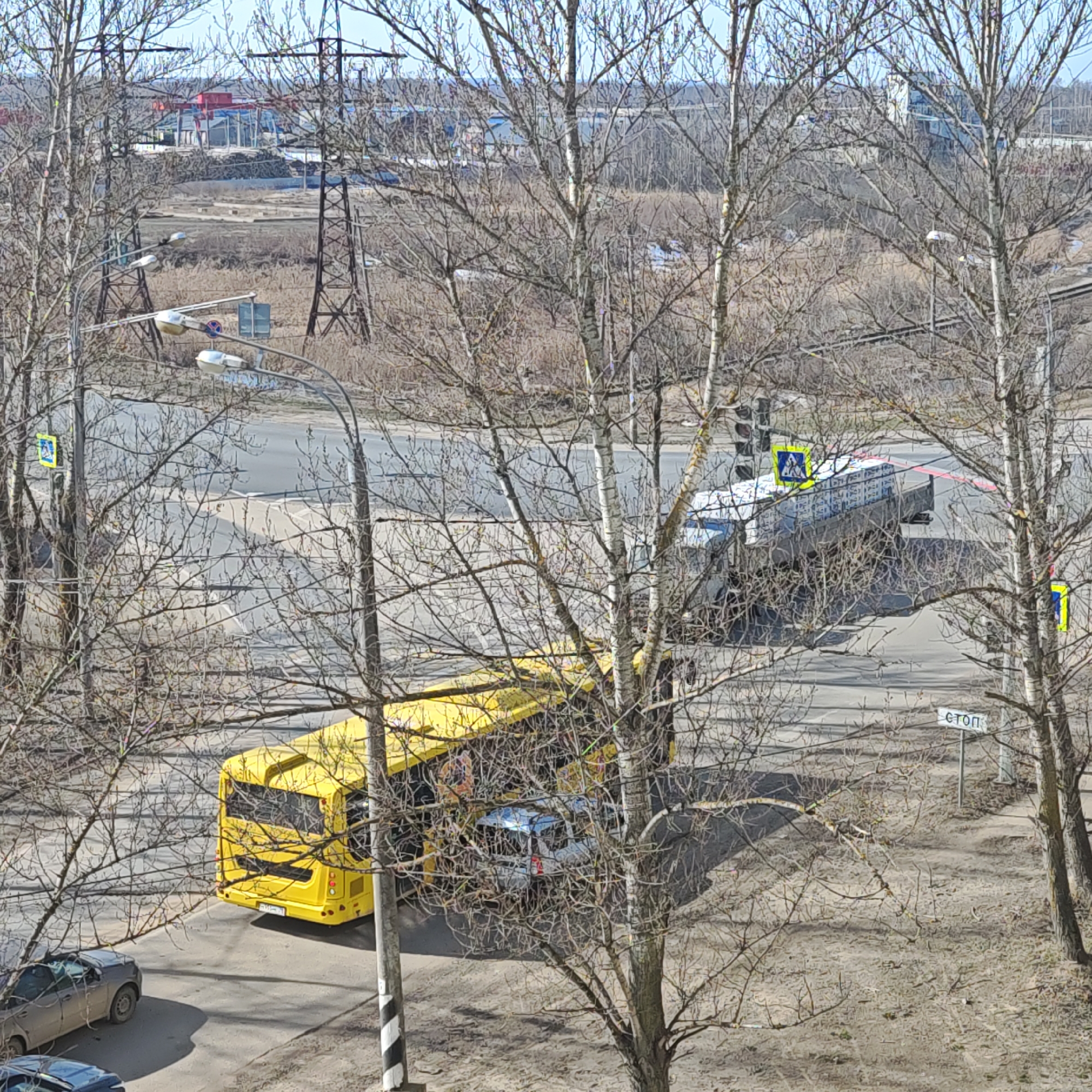 A high-angle shot depicts a street scene in Rostov, Russia. A yellow bus and a light-colored van are near an intersection with a traffic light. A semi-truck is further along the road. Pedestrian crossing signs are visible. A "STOP" sign is present. Trees with bare branches frame the view. Power lines and distant industrial buildings are visible in the background. Several parked cars are visible near the bus.