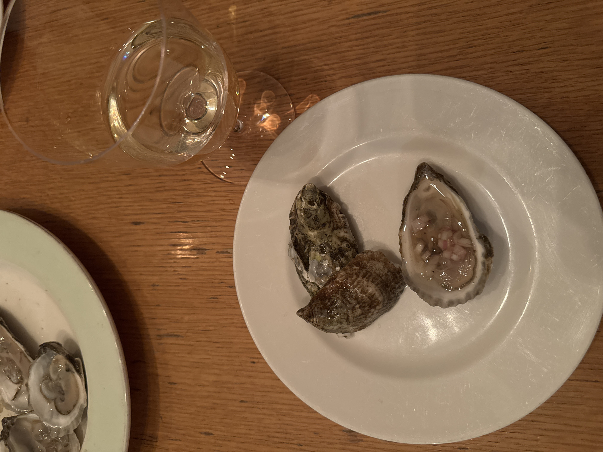 The image displays an overhead view of a dining scene with multiple plates of oysters and a glass of white wine on a wooden table. In the foreground, a white circular plate holds three oysters. One oyster is shucked, containing its meat and a red mignonette dressing. Two other oysters on this plate are partially shelled. In the upper left background, a portion of a light green or off-white plate is visible, containing at least two additional shucked oysters. To the right, a stemmed wine glass is filled with a pale yellow liquid, identified as white wine. The items rest on a polished wooden table with distinct vertical grain patterns. The lighting is dim, suggesting an indoor restaurant or bar setting. The specific location within New York City, United States of America, cannot be determined from the visual information provided. No people are visible in the frame.