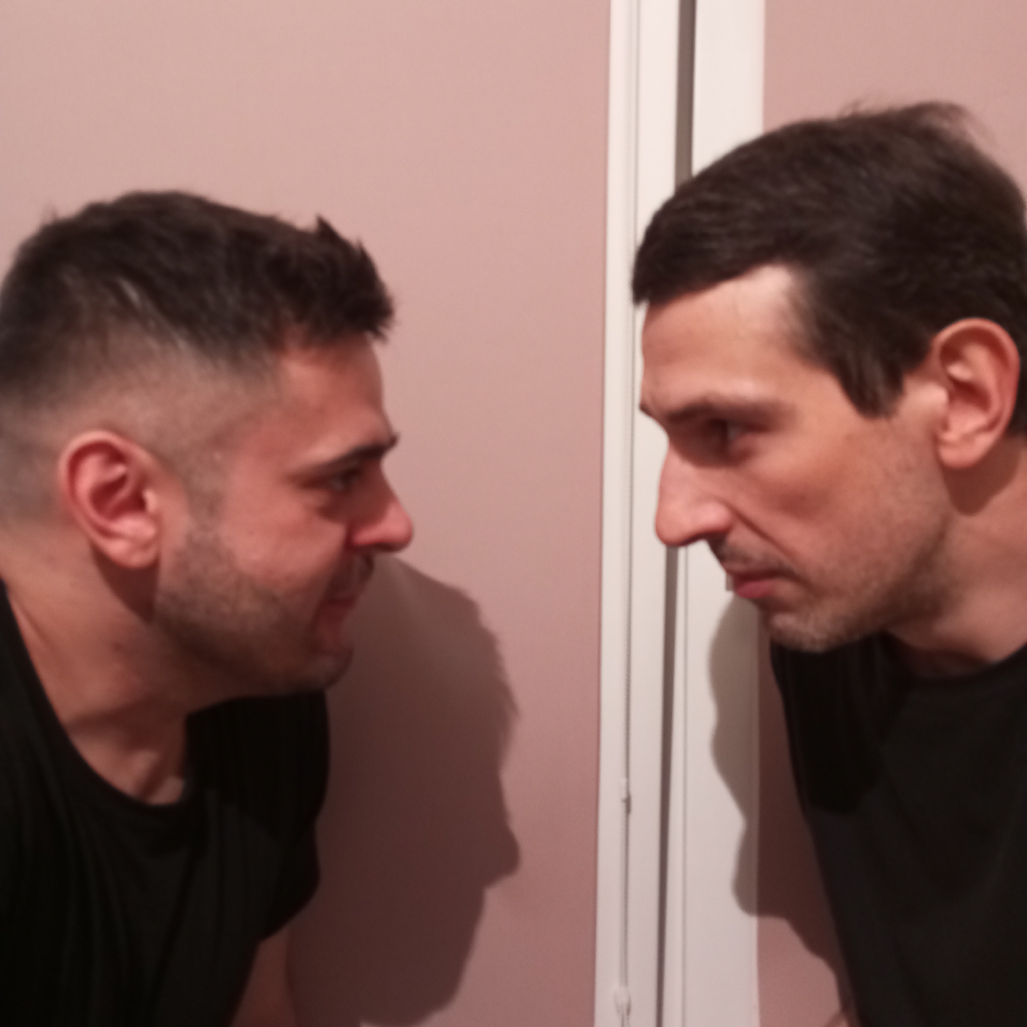 Two men face each other, looking intensely. The man on the left has short dark hair, a beard, and is wearing a black t-shirt. The man on the right has dark hair and is also wearing a black t-shirt. They are positioned against a light pink wall, with a white door frame visible behind the man on the right. The location in Pervomaysky, Russia is not discernible. The image focuses on the facial expressions and close proximity of the two men.