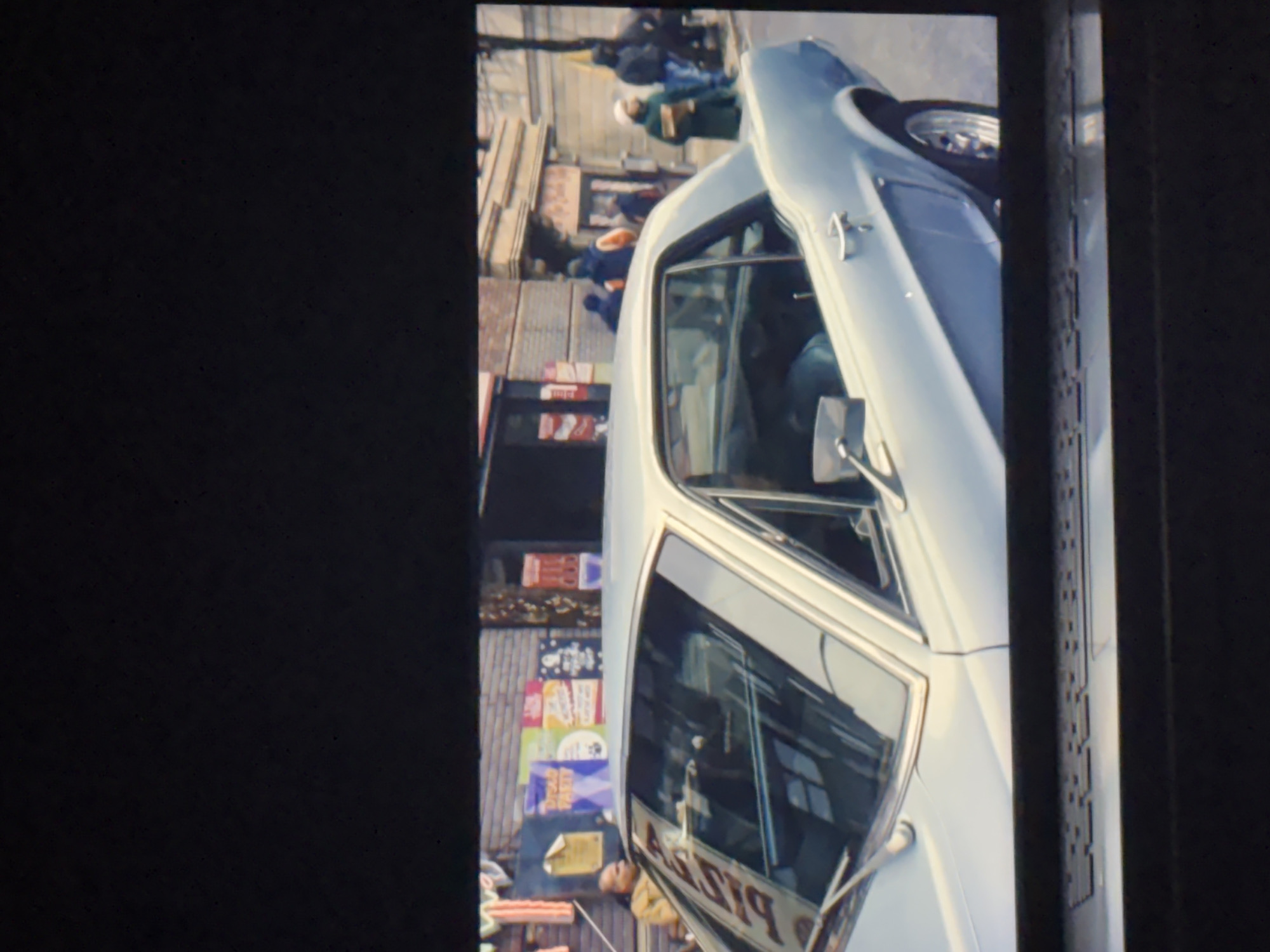 The rotated image captures an urban street scene in Budva, Montenegro, during what appears to be a clear, sunny day. A light-colored (possibly white or very pale blue) classic two-door car with visible chrome accents occupies the right side of the frame, angled towards the viewer. Its front passenger door is slightly ajar, revealing part of the interior. The car features a prominent side mirror and a distinctive wheel design on its visible front tire.
In the background, a busy street is lined with multi-story buildings, likely housing businesses or residences. Several pedestrians are visible walking along the sidewalk, appearing somewhat blurred, suggesting motion or a slightly soft focus. The architecture appears typical of a European city.
A notable detail is the reflection on the car's passenger window, which clearly shows the reversed word "PIZZA," indicating a nearby pizza shop. The buildings in the background are adorned with various signs and posters, some brightly colored. While most text on these signs is indistinct due to distance and resolution, some characters resembling Chinese script are surprisingly visible on one of the signs, alongside other unidentifiable lettering and numbers like "24h."
The scene depicts a typical daytime street environment, filled with the casual activity of city life. No specific event or strong emotions are discernible from the candid snapshot.