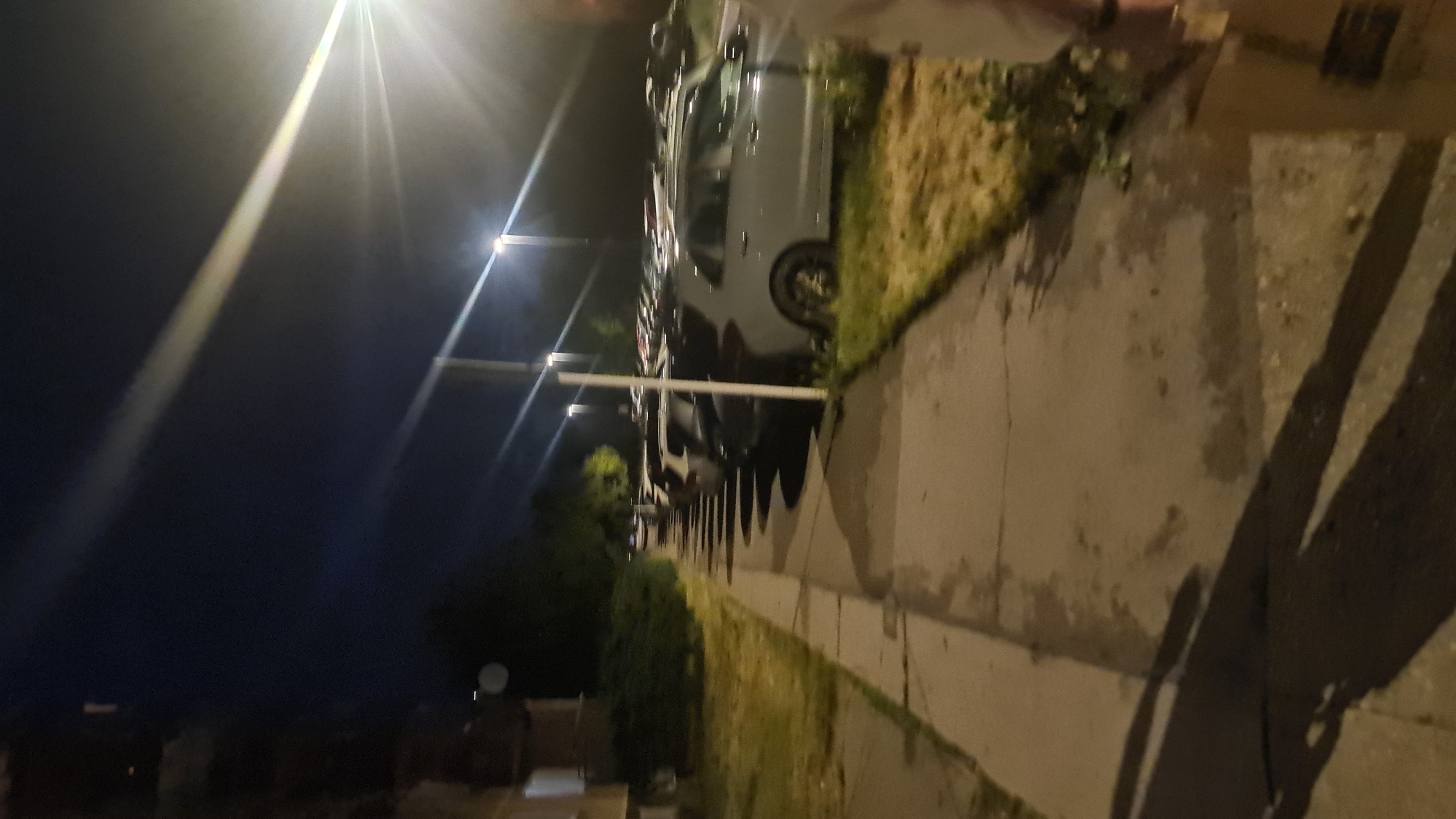 The image shows a row of parked cars along a sidewalk at night. The scene is illuminated by streetlights, casting long shadows on the sidewalk. A grassy area separates the sidewalk from the road. The location appears to be a residential or commercial area with dense foliage.