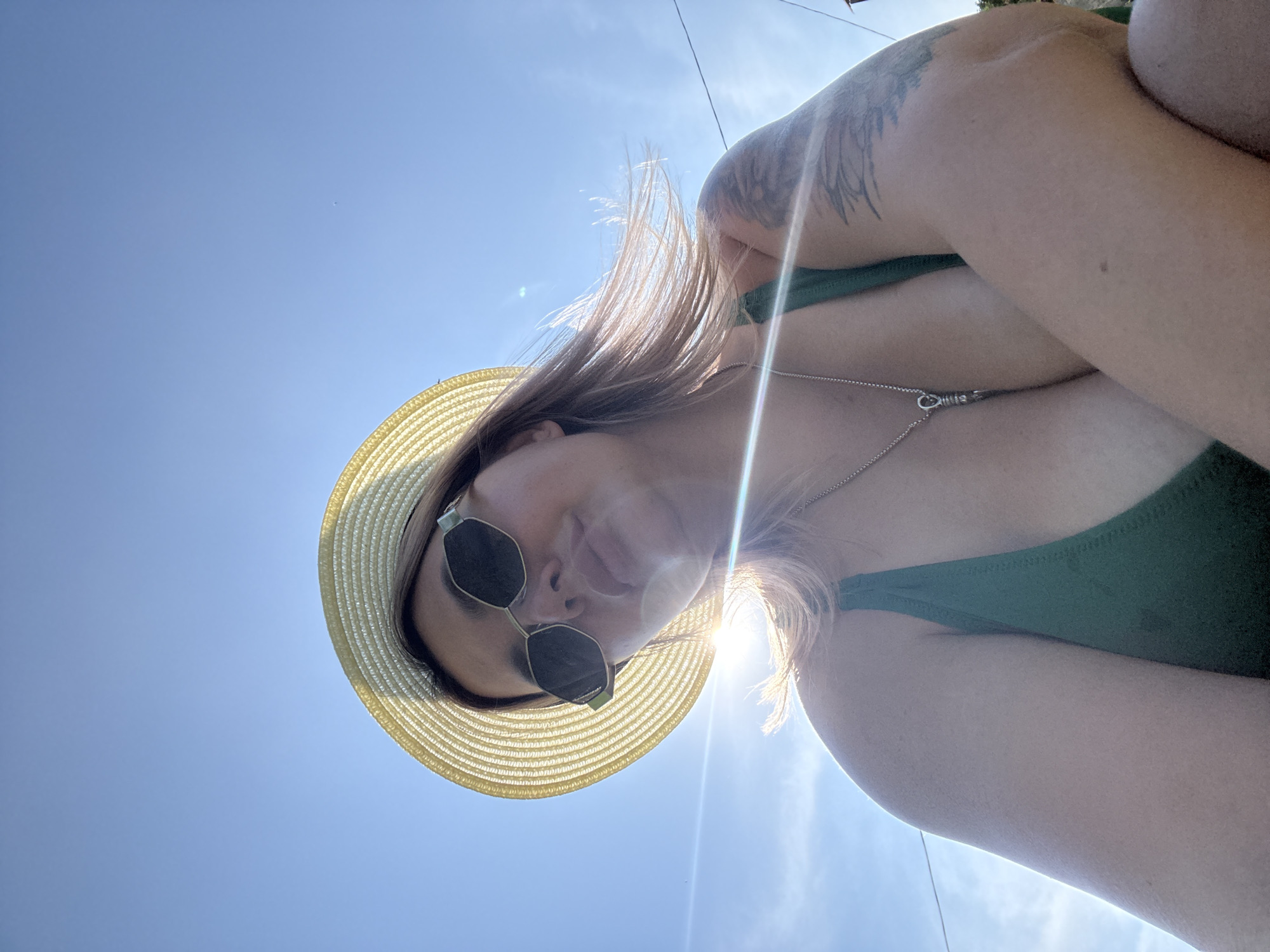 A low-angle, outdoor shot features a person wearing a straw hat and sunglasses, looking down. The person has blonde hair and is wearing a green swimsuit. A silver necklace is visible around their neck, and a tattoo is present on their arm. The bright sun is shining, creating lens flare and casting a glow on the person's hair. The background is a clear blue sky. The image appears to have been taken on a sunny day. There is no visible text in the image.