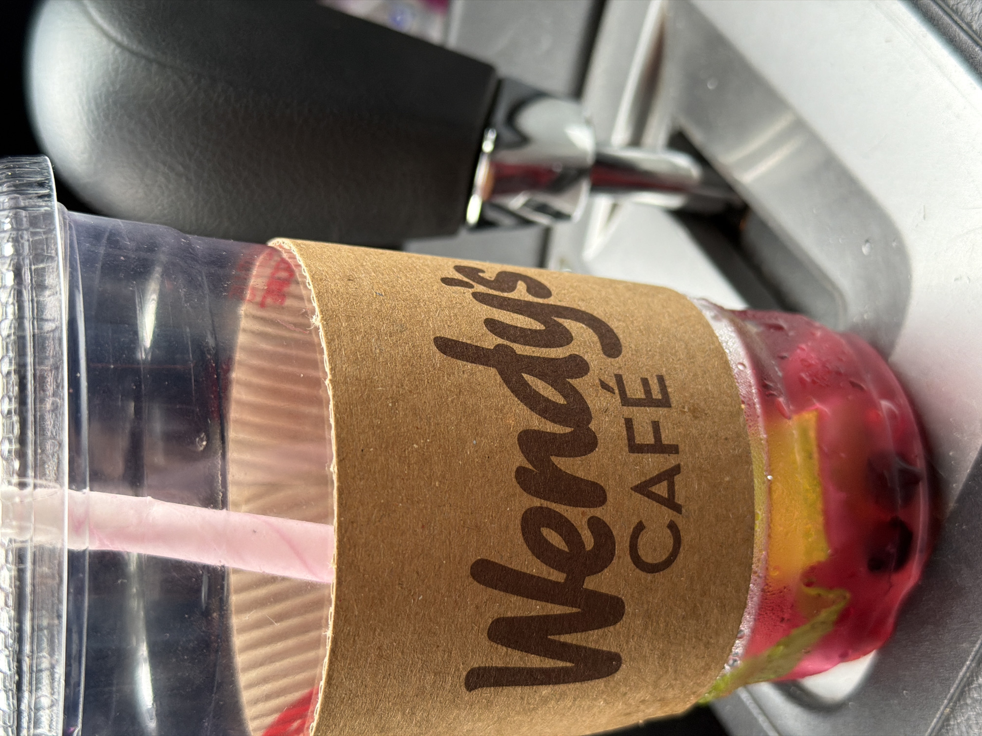 A cup from Wendy's Cafe sits in a car cupholder. The cup is made of clear plastic with a pink straw, and has a brown paper sleeve printed with "Wendy's CAFE" in a brown, stylized font. The contents of the cup are a pink liquid with pieces of fruit, including lemon and what appears to be berries. The cupholder is built into the car's center console, which is light gray. A black gear shift is partially visible above the cup. There is no information available to infer the city of Tbilisi, Georgia from this image.