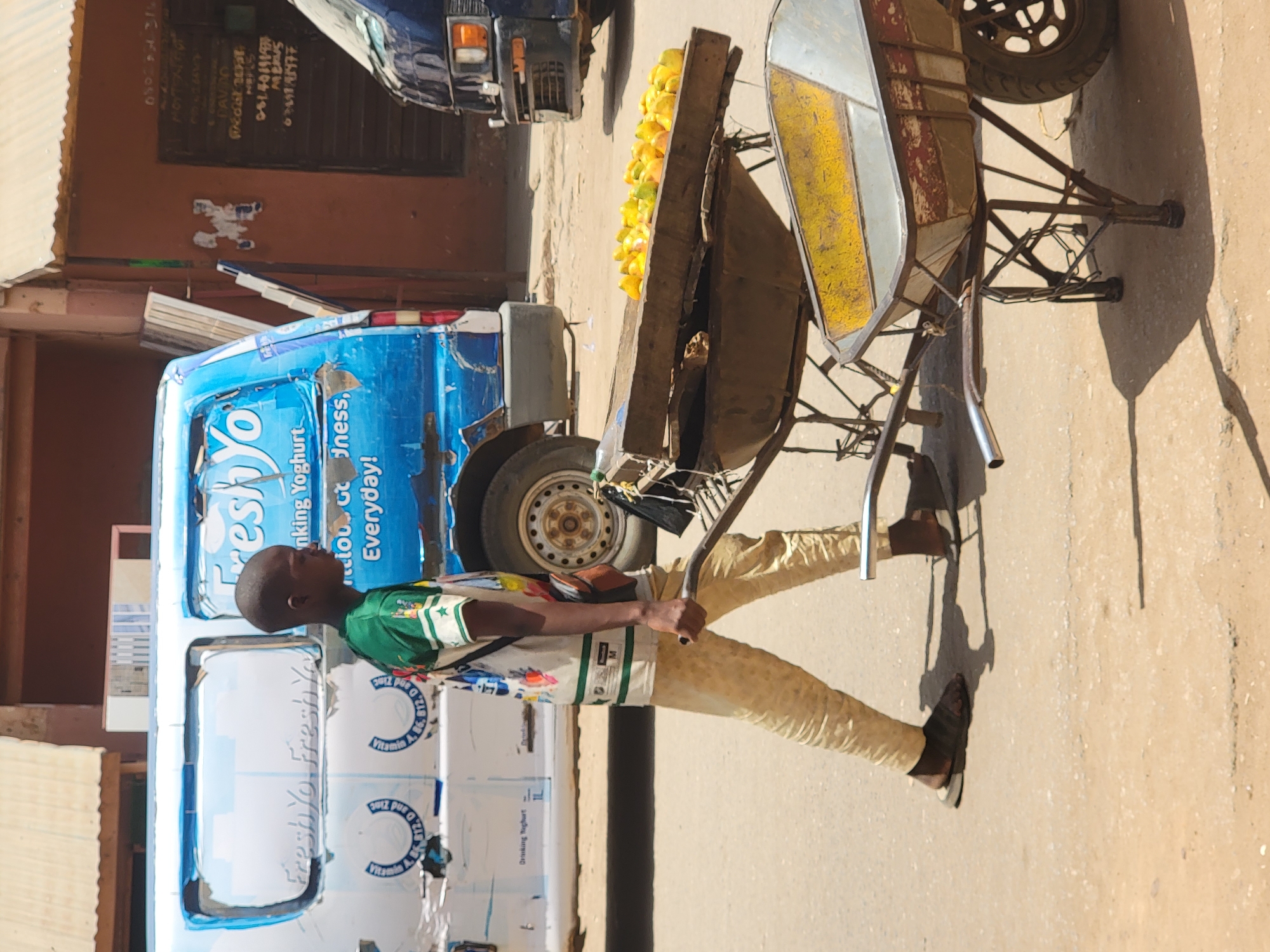 This image captures a vibrant street scene in Maiburiji, Nigeria, under strong midday sun. The central subject is a dark-skinned man, appearing to be a street vendor, standing beside a two-wheeled cart loaded with bright yellow fruits, likely mangoes or oranges. He wears a short-sleeved green and white patterned shirt, light-colored trousers, and sandals, holding the handles of his rustic metal cart.
Behind the vendor is a blue and white commercial van, possibly a mini-bus, branded with "Fresh Yo" logos. Visible text on the van includes "Fresh Yo," "Drinking Yoghurt," and phrases like "dness, Everyday!" and mentions of "Vitamin A, B, E and Zinc." Further in the background, a dark blue vehicle, possibly a truck, is partially visible. The setting is an unpaved road or dirt street, with a reddish-brown building in the background featuring a dark window or door. Faint inscriptions and posters are visible on the building wall, including names like "DAVAO" and "MERUS," along with phone numbers such as "081401919#" and "644742477." The bright sunlight casts sharp shadows on the ground, suggesting it is mid-day or early afternoon with clear weather. The overall impression is a typical bustling street market or vendor scene.