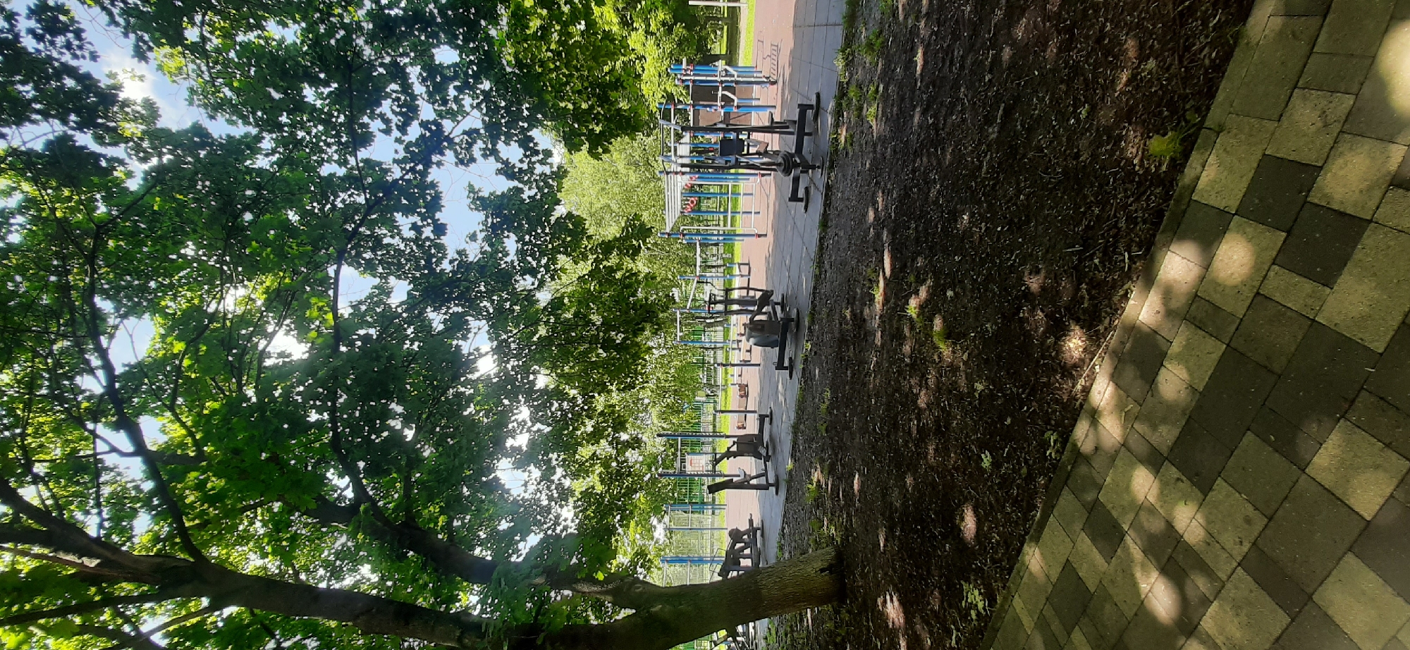 The image shows an outdoor gym with various exercise equipment, including weight machines and bars, set up on a rubberized surface. The area is surrounded by trees, with a pathway made of paving stones in the foreground. The gym appears to be located in a park or recreational area, possibly in the city of Yessentukskaya, Russia, given the context provided. There are no people visible in the image, and the scene is quiet and still. The lighting suggests it is daytime, with sunlight filtering through the leaves of the trees, casting shadows on the ground and pathway.