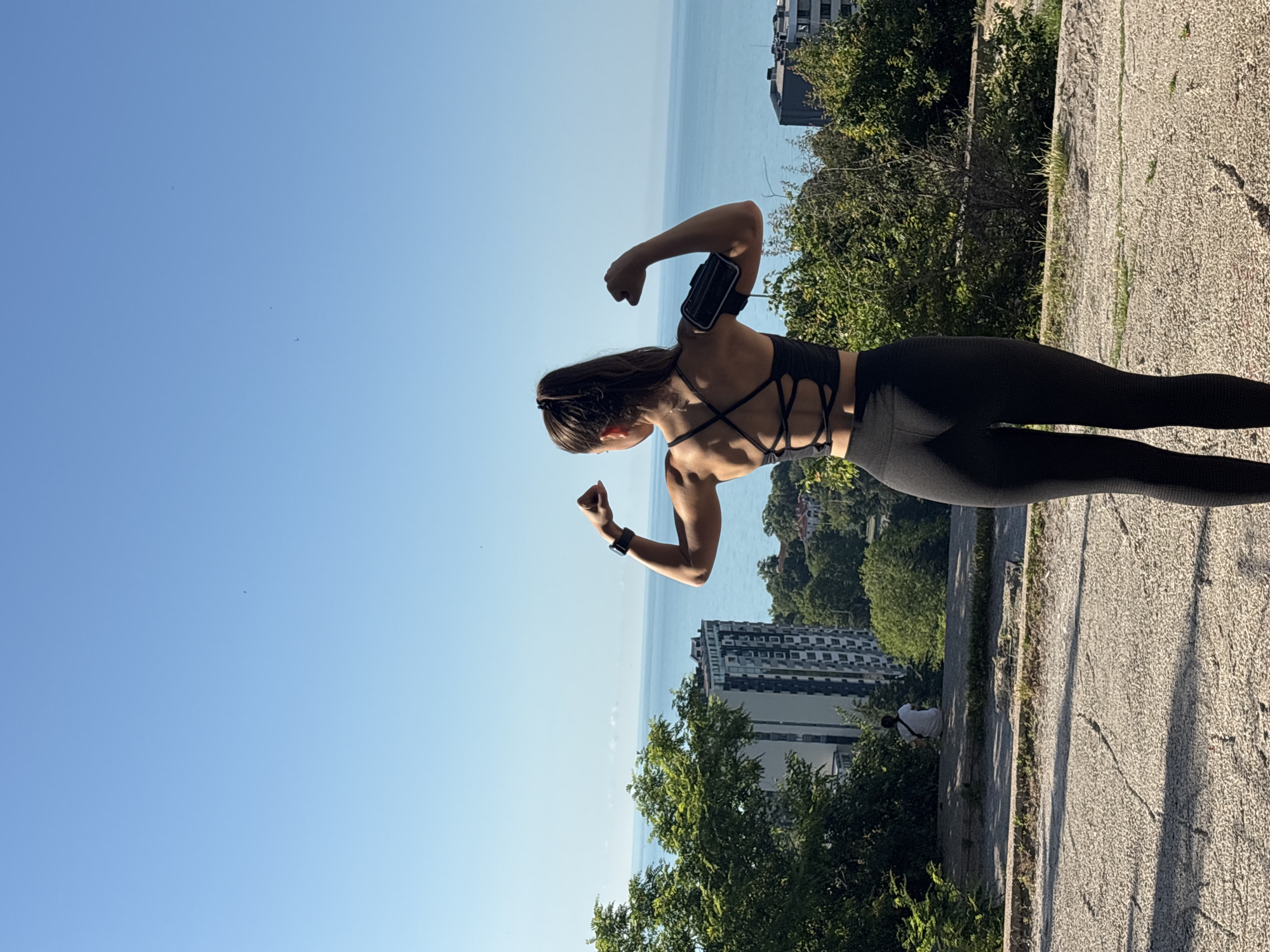 A woman in athletic wear is posing with her back to the camera, flexing her biceps. She is wearing a black sports bra with a crisscross back detail and black leggings. An armband is visible on her left arm. She is standing on a paved surface with a view of the Black Sea and several buildings in the distance. To her left, a person is sitting on a ledge, facing away from the camera. The scene is set against a clear blue sky. This location is likely the central part of Varna, Bulgaria, possibly near the Sea Garden or a viewpoint overlooking the city and the sea.