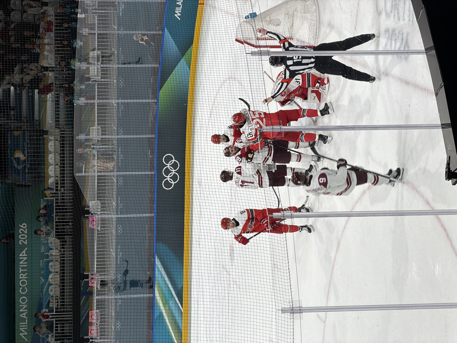 A hockey game shows players in white and red jerseys, engaged in play near the net, officiated by a referee in black and white stripes. Spectators are visible in the background. Olympic rings are emblazoned on the rink barrier. The image is captured from behind the net, providing a side view of the players and the rink. The rink is enclosed by netting.