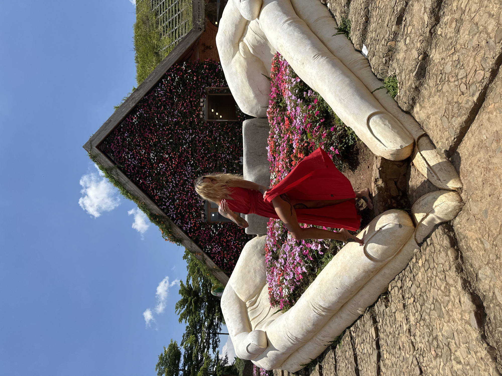 In Cam Ly - Đà Lạt, Vietnam, a woman in a red dress stands in a garden surrounded by colorful flowers. She is positioned between two large, white stone hands that form a heart shape. Behind her, a house with a triangular roof is covered in a dense arrangement of pink and purple flowers, with a green, grassy roof. The sky is blue with scattered white clouds, and trees are visible in the background. The ground is paved with cobblestones.